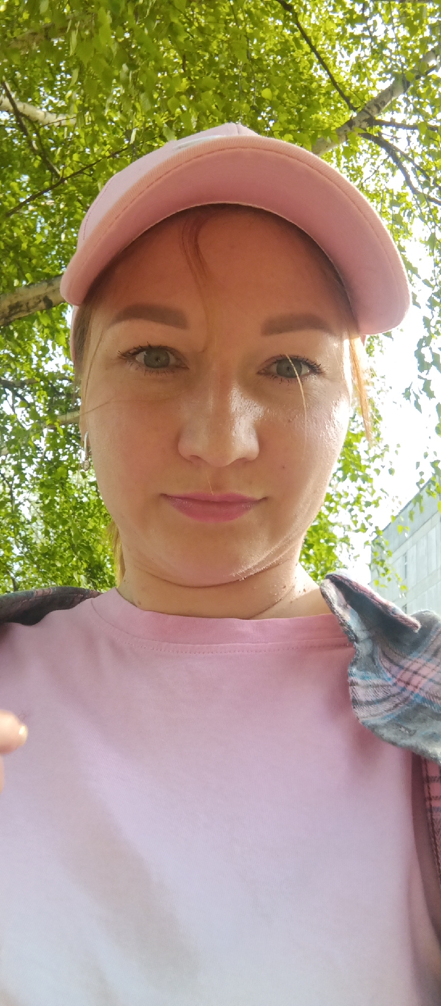 A fair-skinned woman with green eyes and reddish-blonde hair is photographed outdoors in an urban setting. She is wearing a light pink baseball cap, a matching pink t-shirt, and a plaid shirt is draped over her shoulder. Her expression is neutral. Overhead, green leaves from trees fill the upper portion of the frame. The background includes a building, indicating a cityscape. The location is described as Unknown, Unknown.