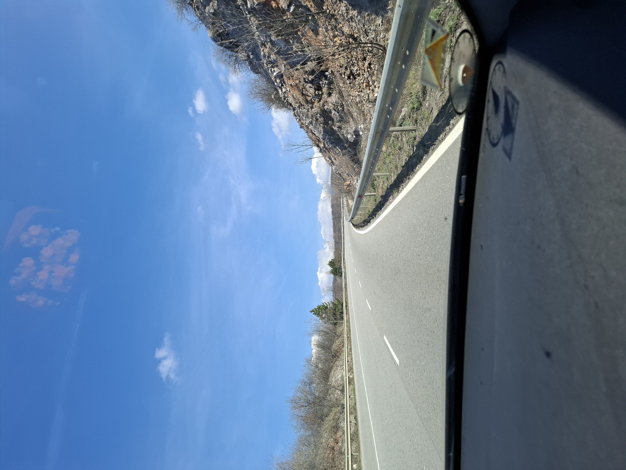 An eye-level, canted, mobile phone photograph, from inside a vehicle, shows an empty, two-lane asphalt road in Bogyovtsi, Bulgaria. To the left of the road is a bright, blue sky with a few small, white clouds, and the road is flanked by an aging guardrail. On the left side of the road, beyond the guardrail, is a rocky hillside. On the right side of the road, beyond the guardrail, is a dry, forested area. In the lower right of the photograph is the inside door frame of the vehicle. The scene is during the day.