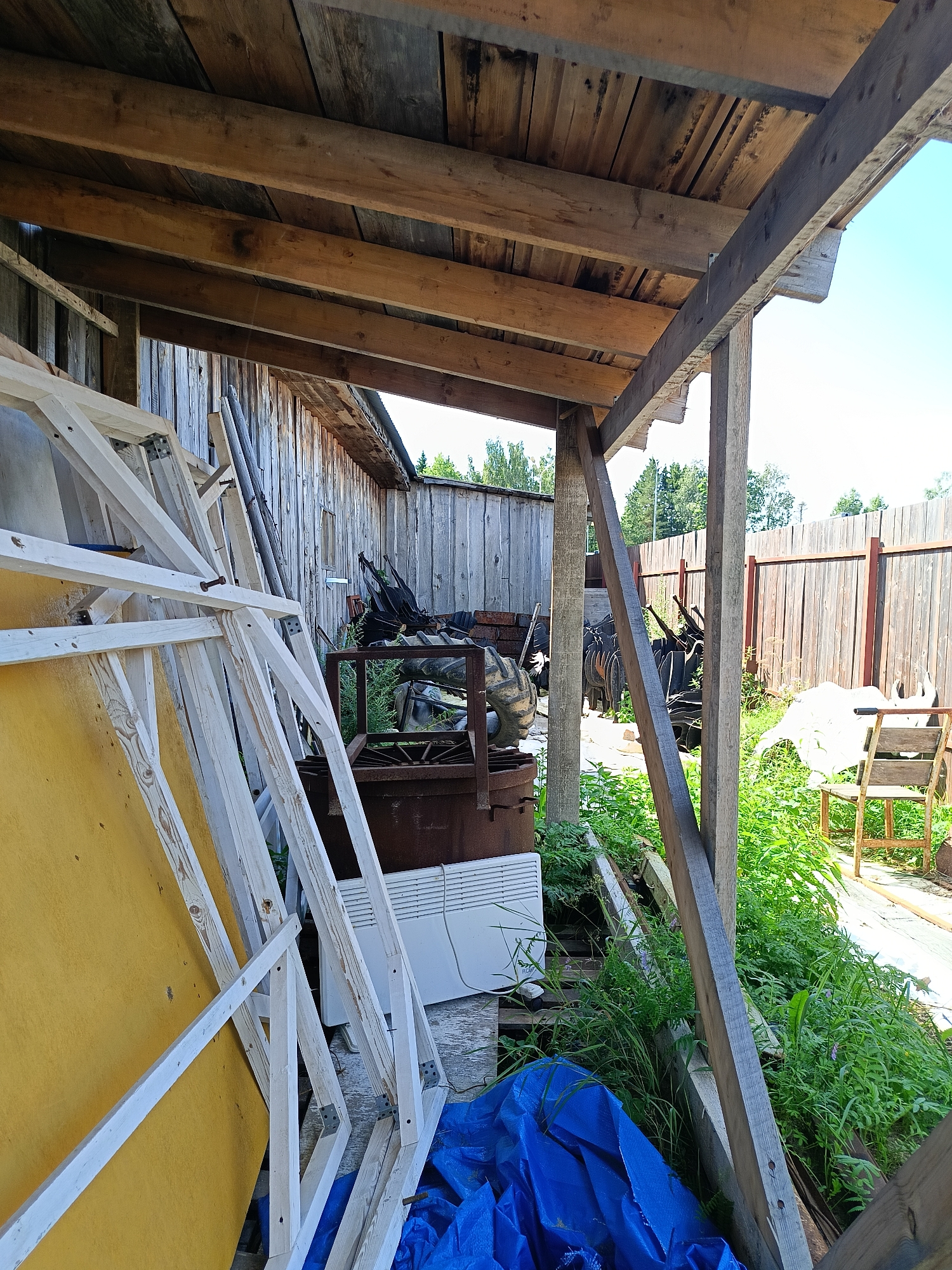 The image depicts an outdoor storage area, possibly a workshop or shed, located in Semenkovo, Russia. The scene is characterized by a rustic, unkempt appearance. In the foreground, to the left, a large yellow panel leans against a structure constructed from white wooden frames. These frames are assembled with metal brackets. Beneath the frames, a bright blue tarp is visible, partially covering the ground and some unseen objects.
The structure's roof is made of weathered wooden planks, supported by rough-hewn wooden beams and posts. The wall of the adjacent building, seen to the left, is constructed of vertical wooden planks.
Further into the scene, various objects are piled up. There's a rusty metal structure, possibly a furnace or grill, with a white heater unit placed in front of it. Behind these items, a collection of agricultural or industrial equipment, including what appear to be tractor parts or plows, is stored. The ground is uneven and covered with weeds and tall green grass, indicating a lack of regular maintenance.
To the right, a wooden fence borders the area. A simple wooden chair with a metal frame stands near the fence, partially obscured by the greenery. The sky is visible in the upper right portion of the image, appearing clear and blue. The overall impression is one of a functional but neglected outdoor space filled with stored materials and equipment. There are no people present in the image.