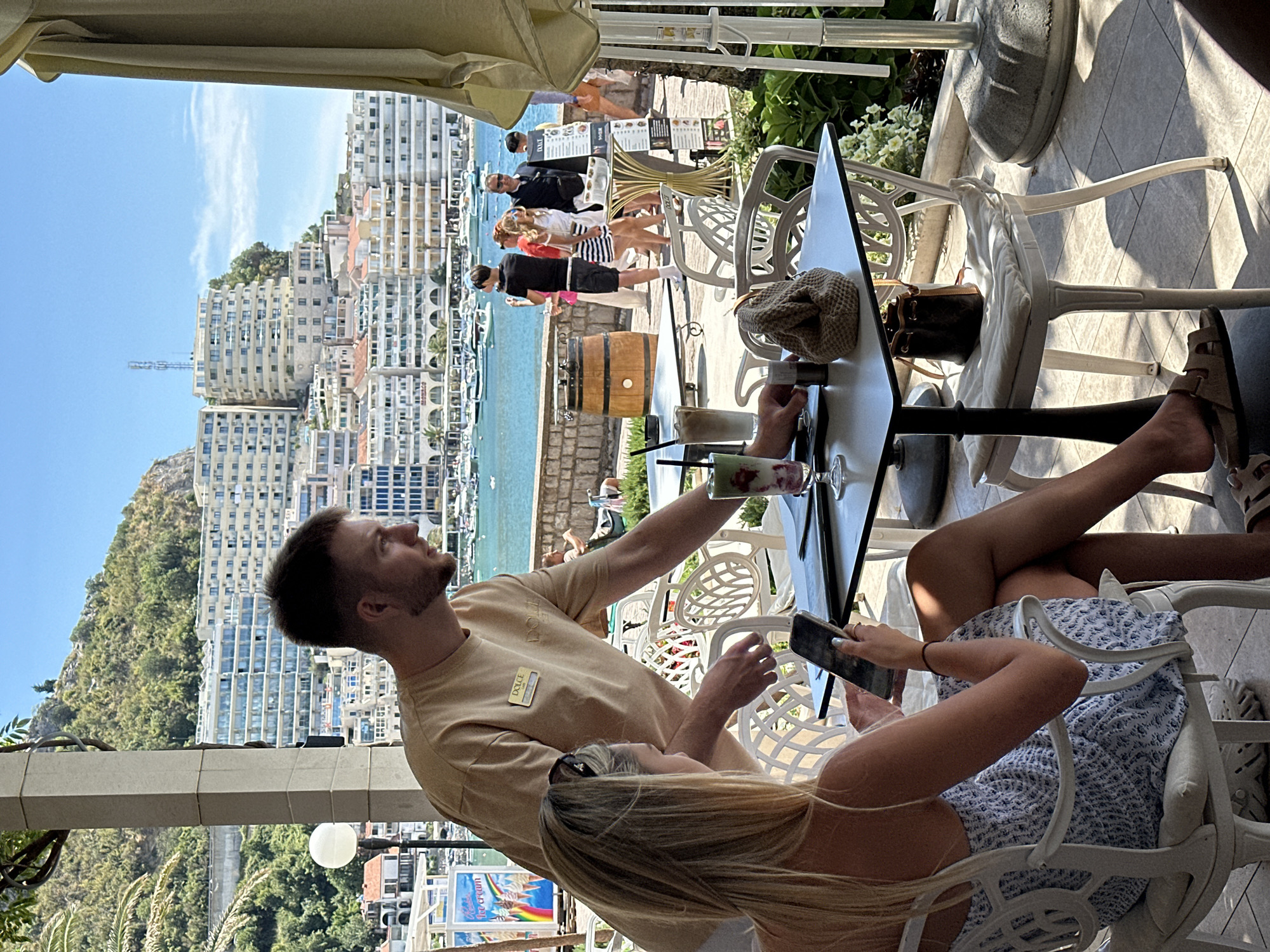 In Rafailovići, Montenegro, a restaurant patron with blonde hair, wearing a white and blue patterned dress, sits at a table and looks at her smartphone. A waiter, identified by a "DOLCE" nametag on his beige t-shirt, approaches her table. On the table, there is a smartphone, a menu, a drink with a straw, and a small knitted bag. In the background, other patrons are visible, as well as buildings and the sea with boats. The setting appears to be an outdoor dining area with umbrellas providing shade.