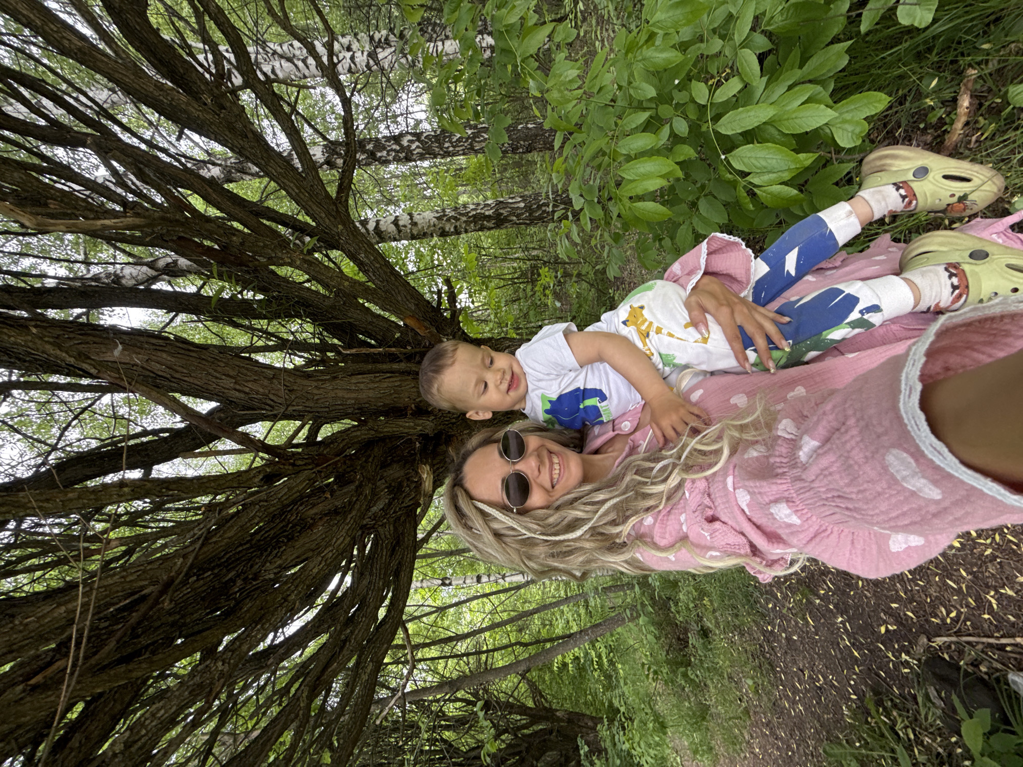 An outdoor portrait shows a woman and a young child, appearing to lie down and look up at the camera, with the image rotated. The woman, with long wavy blonde hair and sunglasses, smiles warmly. She wears a textured pink top or dress with white heart-shaped patterns. A young boy, likely a toddler, rests on her, also smiling and looking slightly upwards. He wears a white t-shirt with a colorful animal design (possibly a giraffe) and blue and white patterned pants. His feet are in white socks and distinctive light green clogs with a dinosaur-like face design.
The setting is a dense forest or wooded area, with numerous tree trunks and branches forming a canopy overhead. Many trees have dark, gnarled bark, while others exhibit the distinct white and black bark of birch trees. Lush green foliage covers the trees and undergrowth. The ground visible beneath them is a mix of dirt, grass, and light-colored plant debris. The bright, natural light indicates daytime. Both subjects appear happy and content, conveying a moment of affection and relaxation in nature. No text is visible in the image.