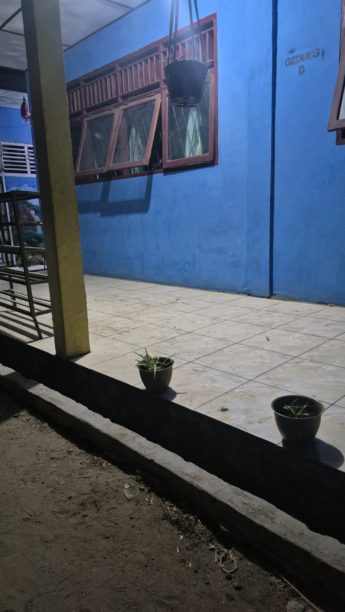 The image depicts an exterior view of a blue building with a window. The window has red wooden frames and is partially open, allowing a view of something inside. A dark hanging basket with a plant is suspended above the window. To the right of the window, the word "GEDUNG" and the letter "D" are visible on the blue wall, suggesting it is building D. In front of the building, a tiled walkway is present. Two potted plants with green shoots are placed on the walkway. To the left of the walkway, there is a yellow pillar and a metal structure with a mural in the background. The foreground of the image shows a dirt area with debris and a concrete edge. The scene appears to be a dimly lit exterior, possibly at dusk or night.