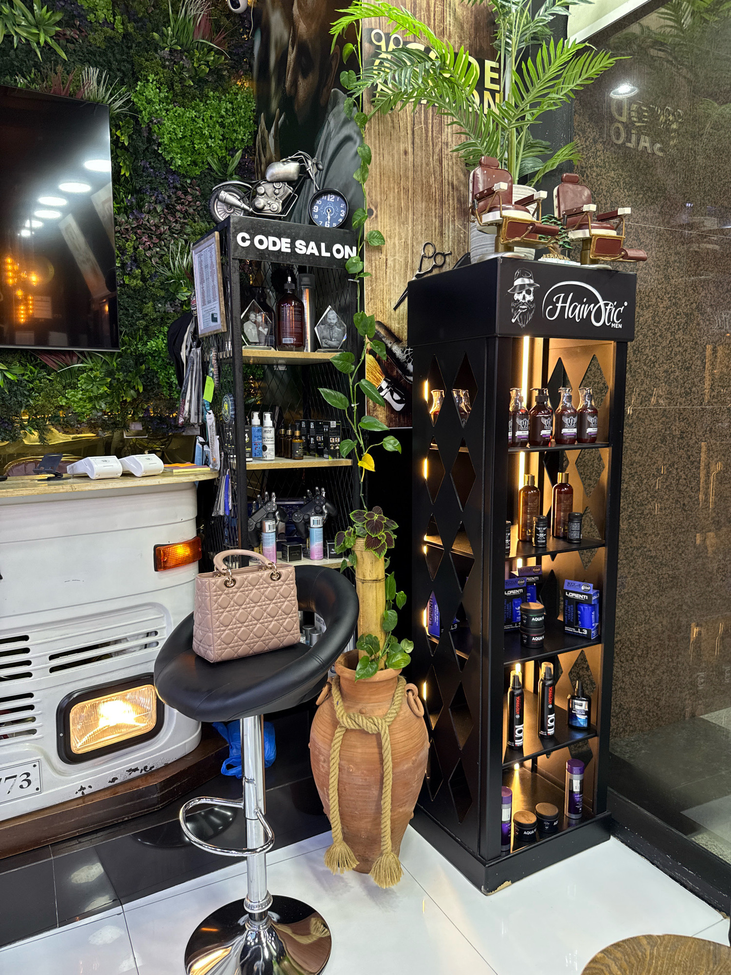 The image depicts an indoor scene, likely a barbershop or salon, with a modern and trendy aesthetic. The left side of the image features a vertical wall covered in lush green artificial plants, creating a natural backdrop. Mounted on this wall is a large flat-screen television. To the right of the plants is a display rack labeled "CODE SALON" with various items, including a miniature motorcycle figurine and a clock.
In the center of the scene, a black bar stool with a pink quilted handbag on its seat is positioned in front of a white vehicle, possibly a custom-built reception desk or vehicle-themed counter, with a prominent orange indicator light. Behind the stool and to the right, a tall, decorative terracotta vase with a rope accent holds more greenery.
The right side of the image is dominated by a black, multi-tiered display shelf filled with men's grooming products and accessories. The top shelf of this display features miniature barber chairs, and the overall display unit is branded with "HairStic MEN." The background shows a textured wall and a glass-paneled entrance or display area. The lighting is warm and ambient, highlighting the products and the textures of the space. There are no people visible in the image. The scene appears to be set in a well-lit, organized retail or service environment.