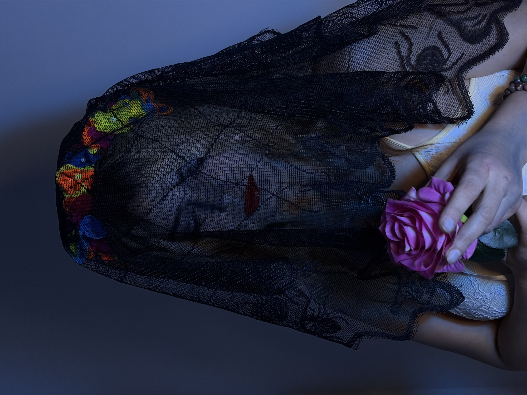 A low-angle, indoor shot shows a person lying down with a black, lacy veil over their face. The background is a dark, cool gray-blue. On the left side of the head are colorful flowers. The person has red makeup over their lips. In the lower right corner, a hand holds a pink rose. A glimpse of a white garment is visible on the right side of the image. The image is rotated counterclockwise. There is no indication of the image being shot in Budva, Montenegro.