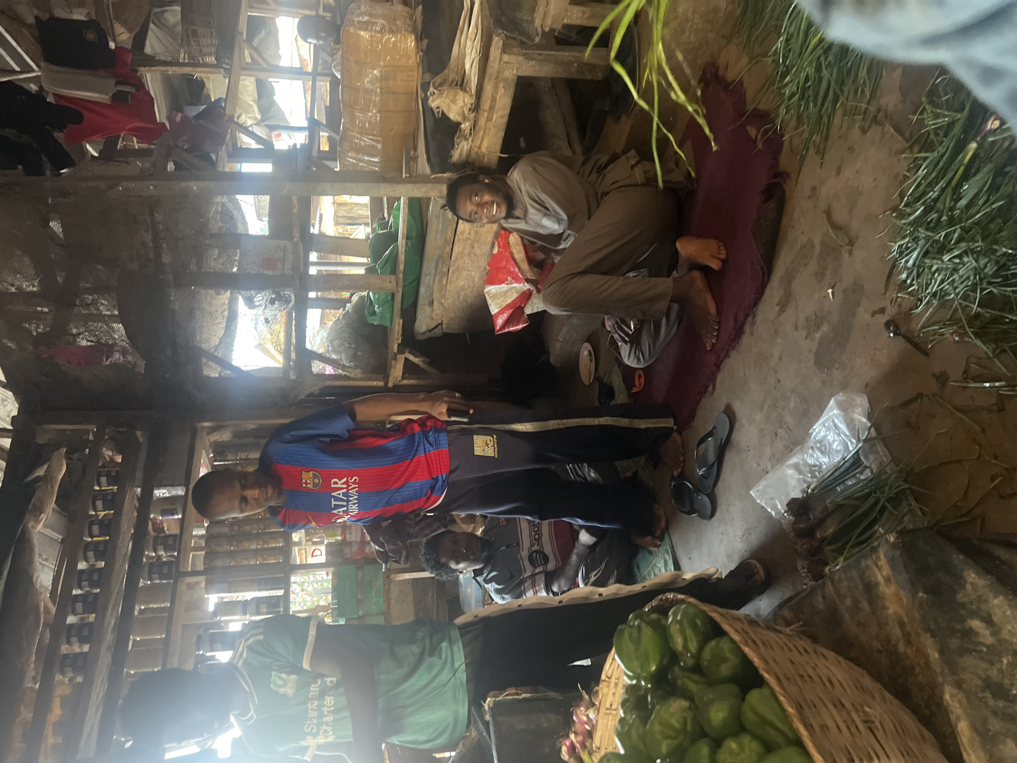 In Oko Amakom, Nigeria, four men are present in a cluttered market stall. Two men stand, one in a green t-shirt and the other in a blue and red striped Barcelona jersey. A third man sits on the floor in a striped shirt, and a fourth man, wearing a light brown outfit, is seated on a purple mat, smiling and holding a red and white item. The stall is filled with shelves of goods, including cans and jars, and various produce like green peppers in a basket and bundles of green onions on the ground. Clothing hangs from the ceiling.