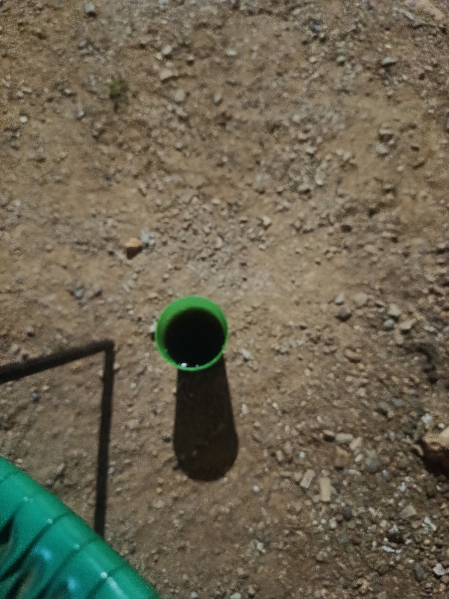 The image shows a patch of rocky, dirt-covered ground. A green object, possibly a tube or hose, is visible in the lower-left corner, casting a shadow. A cylindrical green container filled with a dark liquid is positioned slightly off-center. The location of the image is not identifiable.
