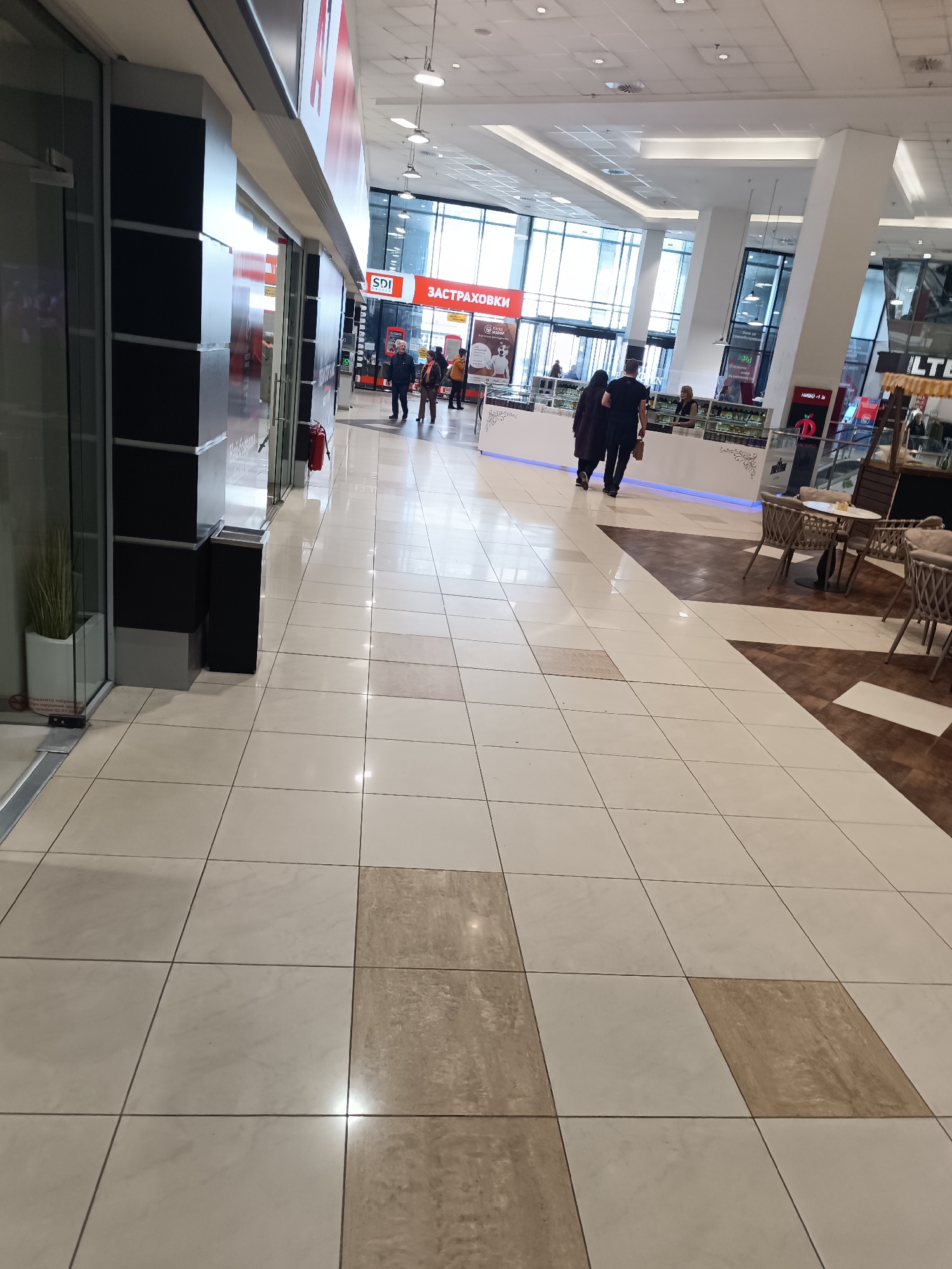 The image shows the interior of a shopping mall in Sofia, Bulgaria. The floor is tiled with a pattern of light and dark beige squares. In the distance, large windows provide natural light. Several people are walking through the mall, including a couple in dark clothing walking away from the camera. Storefronts are visible on the left, with signage in Bulgarian. On the right, there is a café area with tables and chairs. There is an 'SDI Застраховки' insurance booth further down the mall.