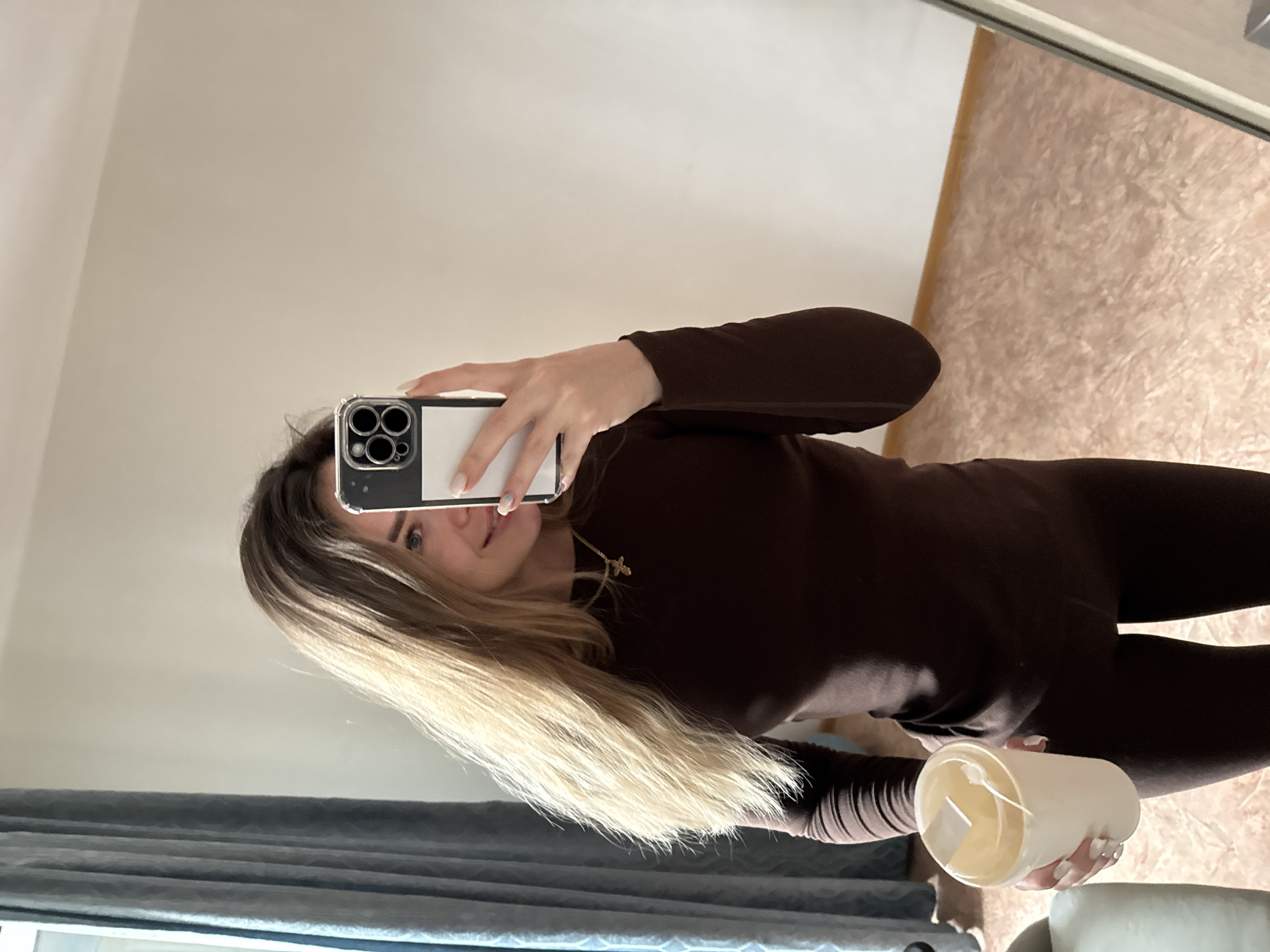 The image captures a mirror selfie of a young woman taken indoors. The woman, with long blonde hair featuring darker roots, is dressed in a dark brown long-sleeved top and matching leggings. Around her neck, a delicate gold cross necklace is visible. She holds an iPhone, identifiable by its triple camera array and a clear-and-white case, positioning it to capture her reflection. Partially visible beneath the phone, her eyes and a slight smile can be seen. Her nails are painted a light color.
In her other hand, she holds a white, cylindrical cup or container. Inside, a light-colored substance resembling wax and a wick are present, suggesting it is a candle. The top edge of the cup appears somewhat irregular.
The setting reflected in the mirror includes a plain, light-colored wall, possibly off-white or beige. The floor has a textured, light brown or pinkish marble-like pattern. In the lower left corner of the reflection, dark grey curtains are visible. The overall lighting is bright and even, indicating an indoor daytime setting or well-lit room. There is no visible text in the image.