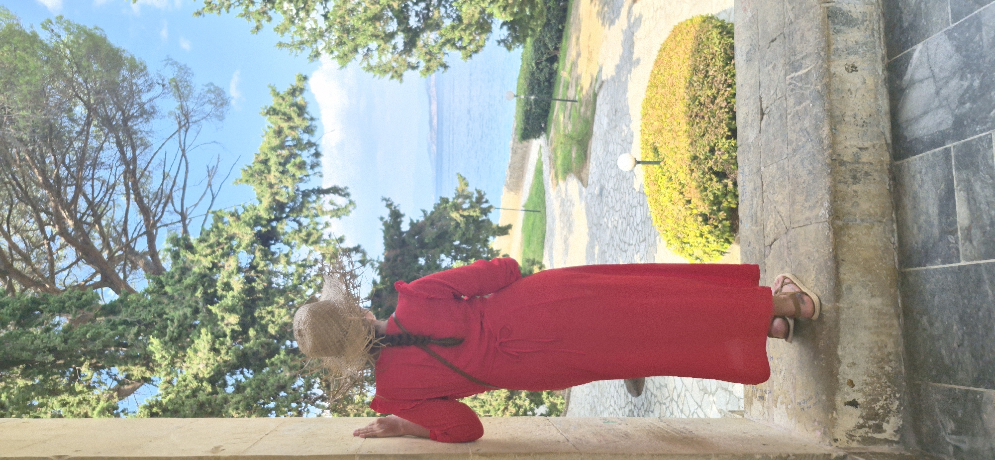 A woman wearing a red dress, a straw hat, and sandals stands at the entrance of a stone building, looking out at the sea. She is leaning against a stone pillar, with her right hand resting on it. Her back is to the camera, and her hair is in a braid. The woman is standing on stone steps, with a pathway and garden visible beyond. The sea is visible in the distance, with hills on the horizon. The scene appears to be a sunny day. The location is the Achilleion Palace, located in the city of Corfu, Greece.