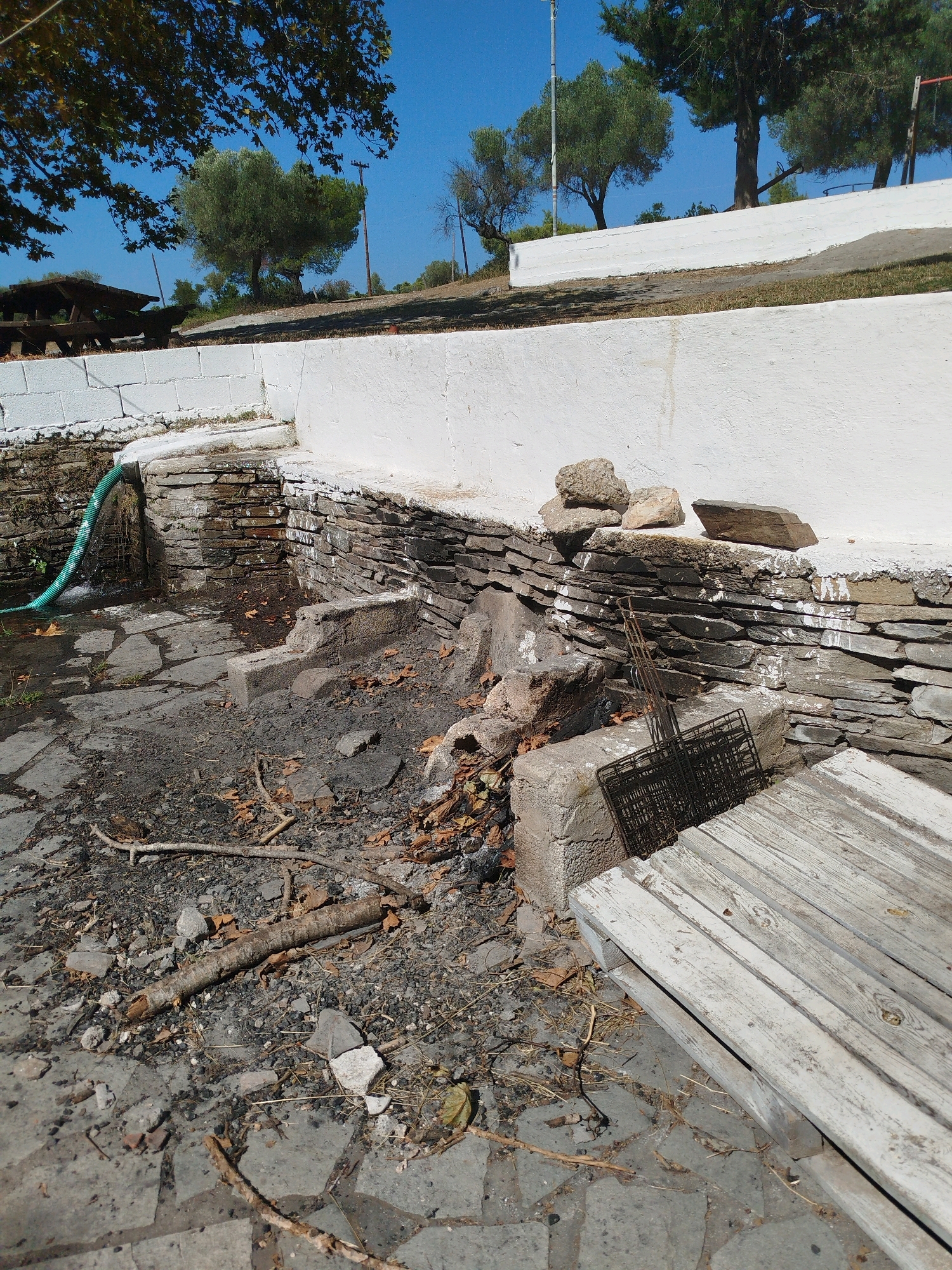 An exterior, ground-level, wide shot captures a rustic, outdoor area with a stone-lined terraced wall. The wall itself is constructed from dark, flat stones layered horizontally, with a white, painted cap. Some of the white paint has chipped and dripped onto the stone. To the left, a green garden hose snakes from behind the wall and empties water onto the stone patio below. The patio is paved with irregularly shaped, large gray flagstones. The ground is covered in dirt, small stones, dry leaves, and scattered twigs, suggesting neglect or recent debris.
In the foreground, on the right, a weathered, light-colored wooden structure, possibly a pallet or bench, rests on the flagstone patio. Behind it, a rusted metal grill rack is propped against a concrete block. The background reveals a clear, bright blue sky above a line of trees, some of which are olive trees, characteristic of the Sithonia Municipal Unit, Greece. A wooden picnic table is visible under the trees in the far left background. The scene appears to be an outdoor seating or gathering area that has fallen into disrepair.