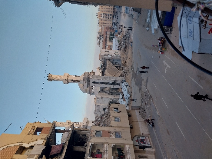 The image, oriented correctly, presents a wide-angle view of a heavily damaged urban landscape, likely after a conflict or disaster, taken from an elevated vantage point. The scene depicts a street flanked by severely damaged buildings, with a prominent mosque standing somewhat intact amidst the destruction.
In the immediate foreground on the left, a multi-story building is significantly destroyed, with large sections of its walls collapsed or missing, exposing interior structures and rebar. Its facade is a mix of light brown and off-white. Further into the left and central background, numerous other buildings are reduced to rubble or show extensive structural damage.
A paved road runs through the center and right side of the frame. It features faded white lane markings, including a solid line along its left edge, dashed lines in the center, and a clear white arrow painted on the right lane, indicating a turn or specific direction. The road itself appears relatively clear, though the areas adjacent to it are filled with debris.
Towards the left-center of the image, behind the foreground damaged structure, a light-colored mosque stands. Its tall, ornate minaret is largely intact and prominently visible, crowned with decorative elements. Part of the mosque's dome can also be seen. The area immediately surrounding the mosque also appears to have sustained damage.
On the right side of the street, there are at least two large white tents or temporary structures. One of these tents has visible text or graphics in a non-Latin script, likely Arabic, which is difficult to decipher clearly. A thick black cable or pipe arcs into the frame from the top right corner, and a thinner, dotted line (possibly a power or communication line) stretches across the clear sky, passing above the mosque.
Few people are visible, contributing to a sense of desolation. Two figures in dark clothing are walking on the middle-right side of the street. Further down the street to the right, a solitary dark silhouette of a person is walking away from the viewer. Near the bottom-left, a small group of approximately three to four people are gathered or moving. Other distant figures can be discerned further down the street and near the tents.
The sky is a clear, light blue, suggesting either early morning or late afternoon, as indicated by the soft lighting and the presence of some shadows, but no harsh direct sunlight. The overall atmosphere is somber, marked by widespread devastation, with the standing mosque minaret serving as a focal point of resilience or endurance amidst the ruin. On a building facade in the bottom-left, a sign or banner is visible, depicting a red pickup truck with text underneath in a non-Latin script (likely Arabic).