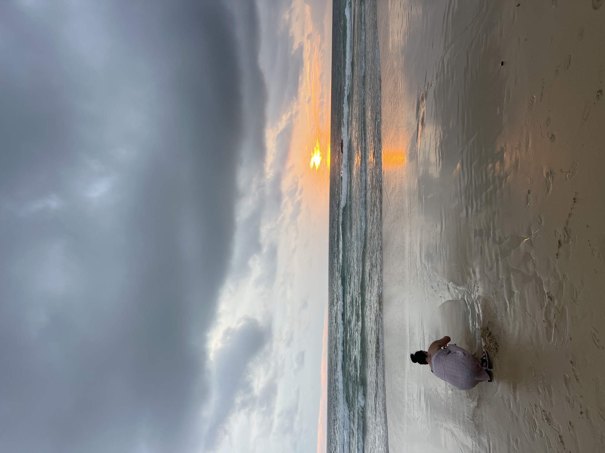 The image shows a person crouching on a sandy beach in Choeng Thale, Thailand, facing the ocean during what appears to be sunset. The sky is filled with heavy, dark gray clouds, but there is a break in the clouds towards the horizon where the sun is setting, casting a bright orange and yellow glow. The sun's reflection is visible on the wet sand and the surface of the ocean. The waves are moderately sized, with white foam breaking on the shore. The person is wearing a light-colored dress with a subtle pattern and is positioned in the lower-left quadrant of the image, looking down towards the sand. The beach shows evidence of water receding, creating a reflective surface on the sand. Footprints are visible in the sand in the foreground.