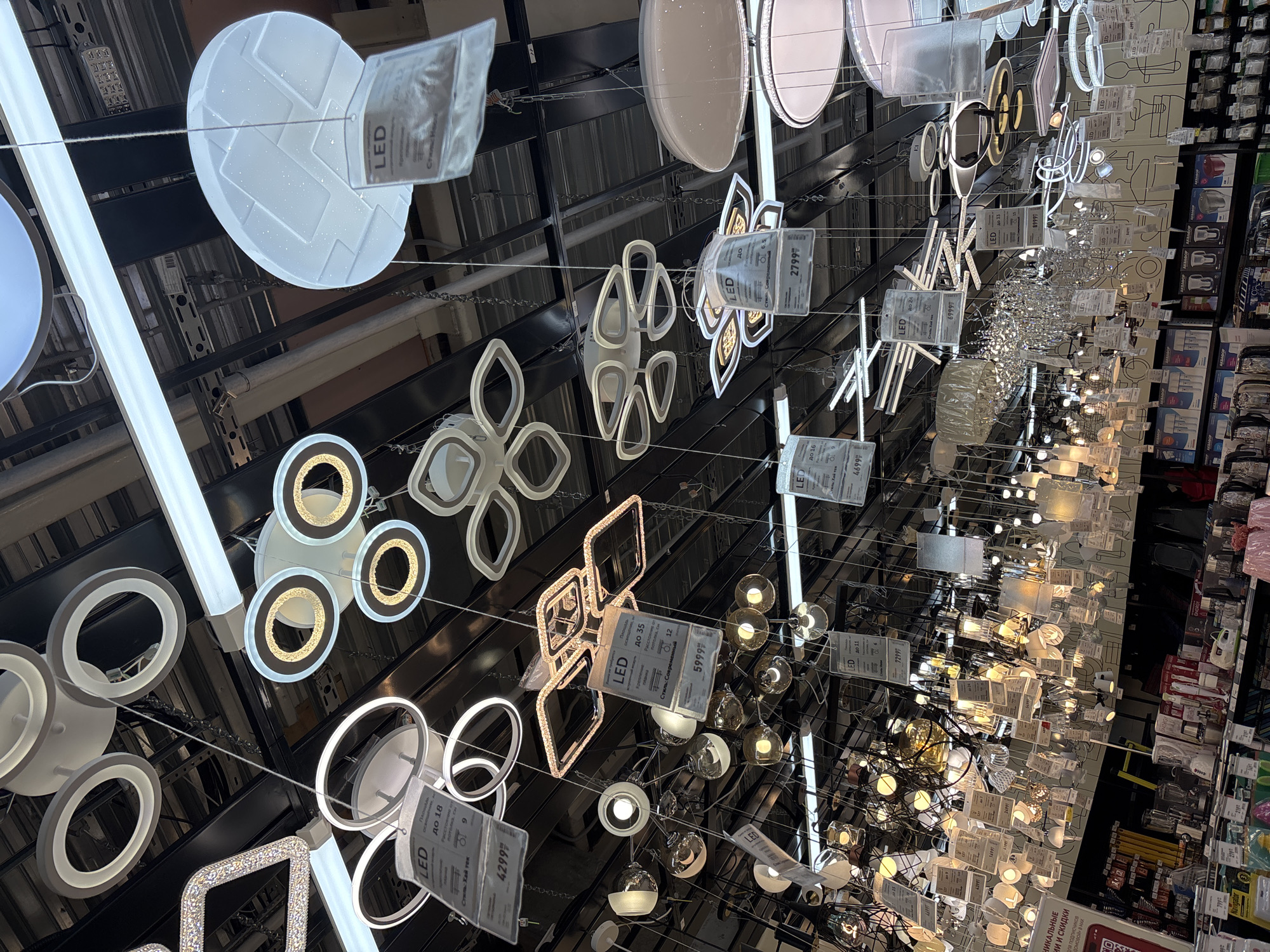 The image captures a vibrant display of numerous LED light fixtures hanging from a ceiling in what appears to be a retail store, likely a home improvement or lighting specialty shop. The lights are varied in design, including modern geometric shapes, circular and oval forms, abstract floral patterns, linear tubes, and some traditional crystal-like chandeliers. Many of the fixtures are illuminated, showcasing their glow and design.
The ceiling infrastructure, consisting of metal beams and tracks, is visible above and between the hanging lights. Price tags and information labels, primarily in Russian, are attached to many of the light fixtures. These labels indicate "LED" technology and feature various prices such as "4299", "5999", "2799", "6999", and "8999", presumably in Rubles. Some tags also include descriptive text like "Стиль: Хай тек" (Style: Hi-tech) and "Стиль: Соврем" (Style: Modern), along with numerical ratings like "до 18", "до 35", "до 16", "до 11", "до 20", and "до 24", which might refer to wattage or coverage area. Partially visible text mentions "НИКАЛЬНЫЕ ИИ И СКИДКИ" (Unique deals and discounts) and "для подписчиков ДОМОВОЙ» В МАХ" (for subscribers of "DOMOVOY" in MAX), suggesting promotional offers related to a store or magazine named "DOMOVOY". In the background, shelves stocked with various household appliances and products are visible, further confirming a retail environment. The lighting conditions are bright and artificial, typical of an indoor commercial setting. The context of the visible Russian text points to the location being in Russia.