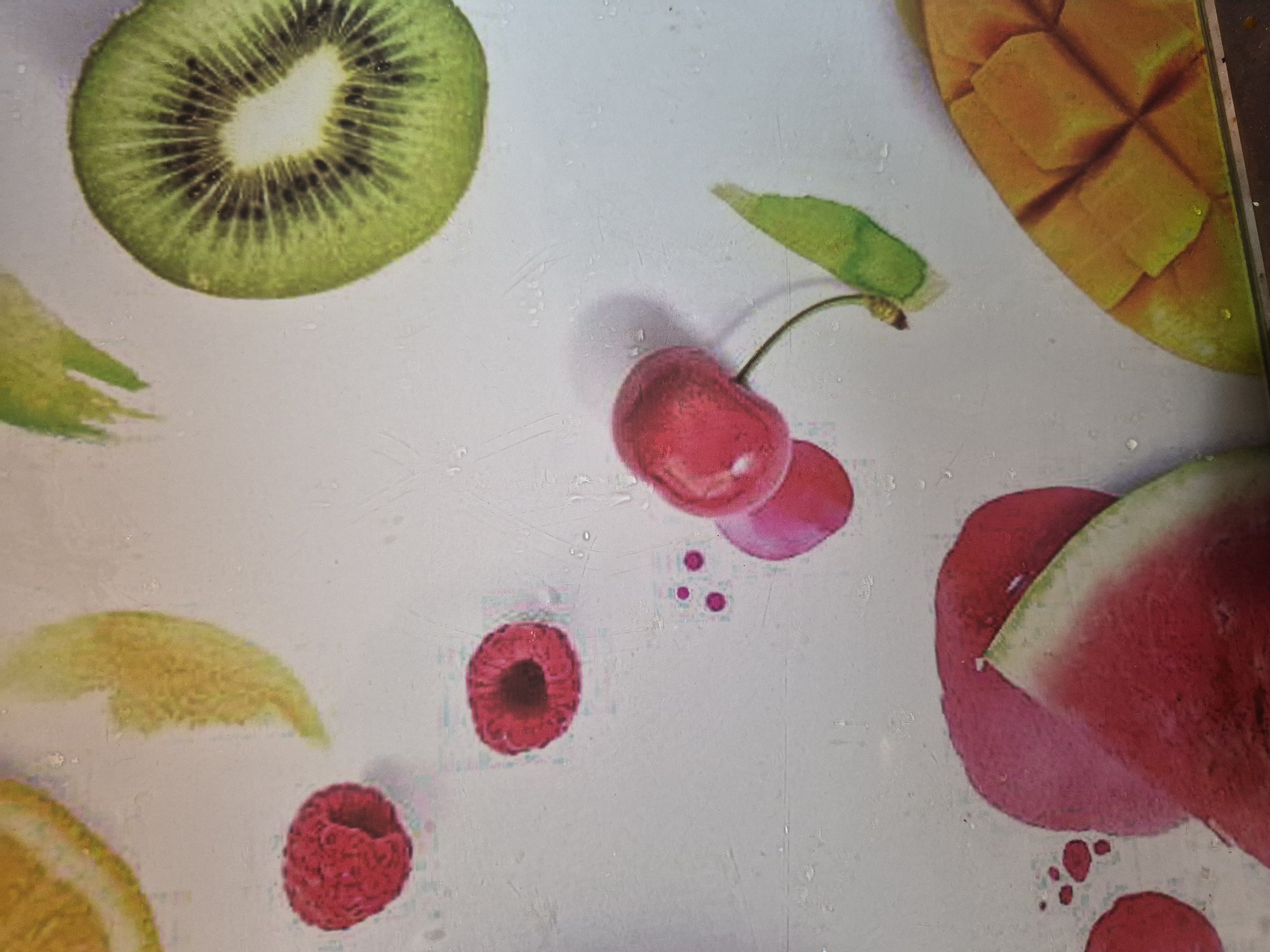 This image displays a brightly lit flat surface, likely a kitchen counter or cutting board, adorned with various colorful fruits. The surface is light grey or off-white and features numerous small water droplets, suggesting a fresh or recently rinsed appearance.
Prominently featured are:
- A cross-section of a kiwi fruit in the upper left, showcasing its vibrant green flesh, dark seeds, and white core.
- A pair of red cherries with a green stem and leaf in the upper-center, casting a soft shadow.
- A cubed section of a ripe mango in the upper-right corner, displaying its rich orange-yellow hue.
- A single, bright red raspberry (or similar berry) in the lower-center, with its textured surface clearly visible. There are also smaller red splatters or liquid droplets nearby.
- A triangular slice of watermelon in the lower-right, revealing its deep red flesh and green rind. This fruit is surrounded by what appears to be red juice or liquid stains.
- A partial slice of a citrus fruit, possibly a lemon or orange, in the lower-left, showing its yellow pulp and white pith. Another red berry is partially visible further down in the lower-left corner.
The arrangement of fruits suggests a still-life composition rather than an ongoing activity. There are no people visible, nor any discernible text. The image evokes a sense of freshness and natural vibrancy.
