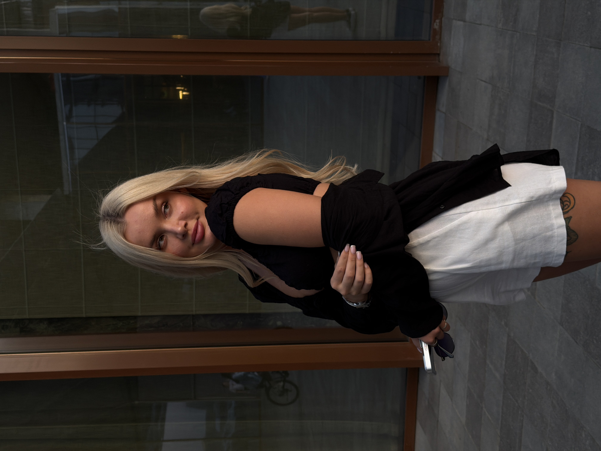 A young woman with long blonde hair is captured in a half-body portrait, standing outdoors. She looks directly at the viewer with a subtle smile, her head tilted slightly, showcasing her fair skin and full lips. She is wearing a black long-sleeved top or open jacket over a white undershirt or dress, and a white skirt or shorts. A dark tattoo with a circular pattern is visible on her right thigh. Her left hand holds a pair of dark sunglasses and a silver-colored object, possibly a phone or phone case, resting against her hip.
The background consists of large, dark-tinted glass windows with dark brown frames, reflecting elements of the surroundings. A faint reflection of a person, possibly the subject herself, is visible in the upper window pane, while a bicycle is dimly reflected in the lower section. The ground beneath her is paved with dark grey tiles arranged in a diagonal pattern. The lighting suggests it is either late afternoon or early evening, creating a subdued and soft atmosphere. The scene evokes an urban or architectural setting, likely a modern building exterior. There is no visible text in the image.