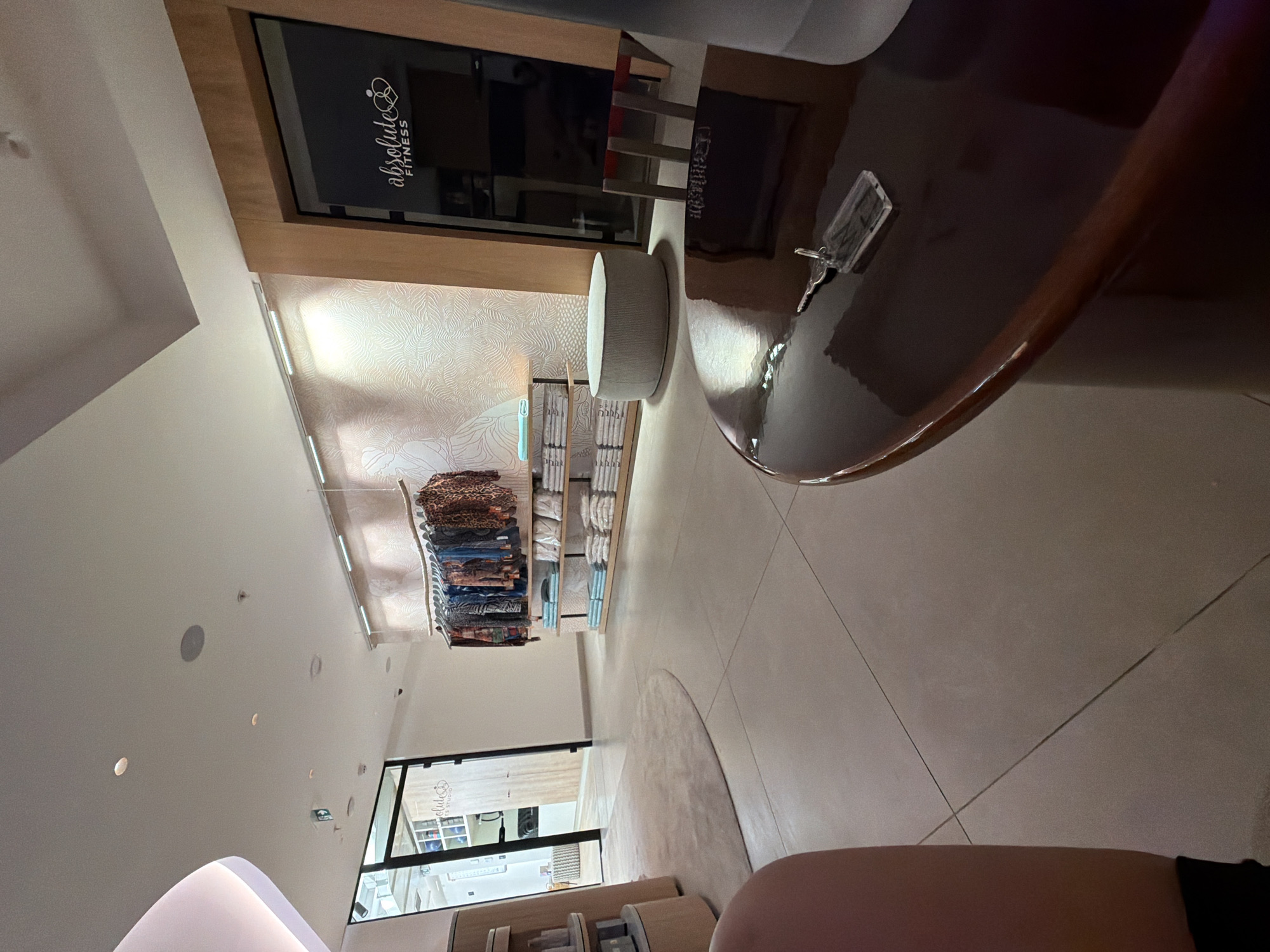 The image depicts the interior of a retail space, likely a boutique or studio, in Budva, Montenegro. The scene is brightly lit, with a combination of recessed ceiling lights and strip lighting illuminating a textured, light-colored wall with a subtle organic pattern. In the foreground, a dark, highly polished circular table occupies the lower right portion of the frame, reflecting the ambient light. A set of keys with a clear rectangular tag rests on the table's surface.
Further into the space, a clothing rack displays a variety of garments, featuring vibrant patterns and colors. Below the clothing rack, several shelves are neatly stacked with folded white towels or linens. To the left of the clothing display, a light-colored, round ottoman-style stool sits on the floor.
An open doorway, framed by wooden trim, leads into an adjacent area. Through this doorway, several shelves are visible, holding various items, and a portion of a sign with the logo "absolute FITNESS STUDIO" can be discerned. The floor throughout the visible area is a smooth, light-colored tiled surface. The overall impression is one of a clean, modern, and well-organized retail environment.