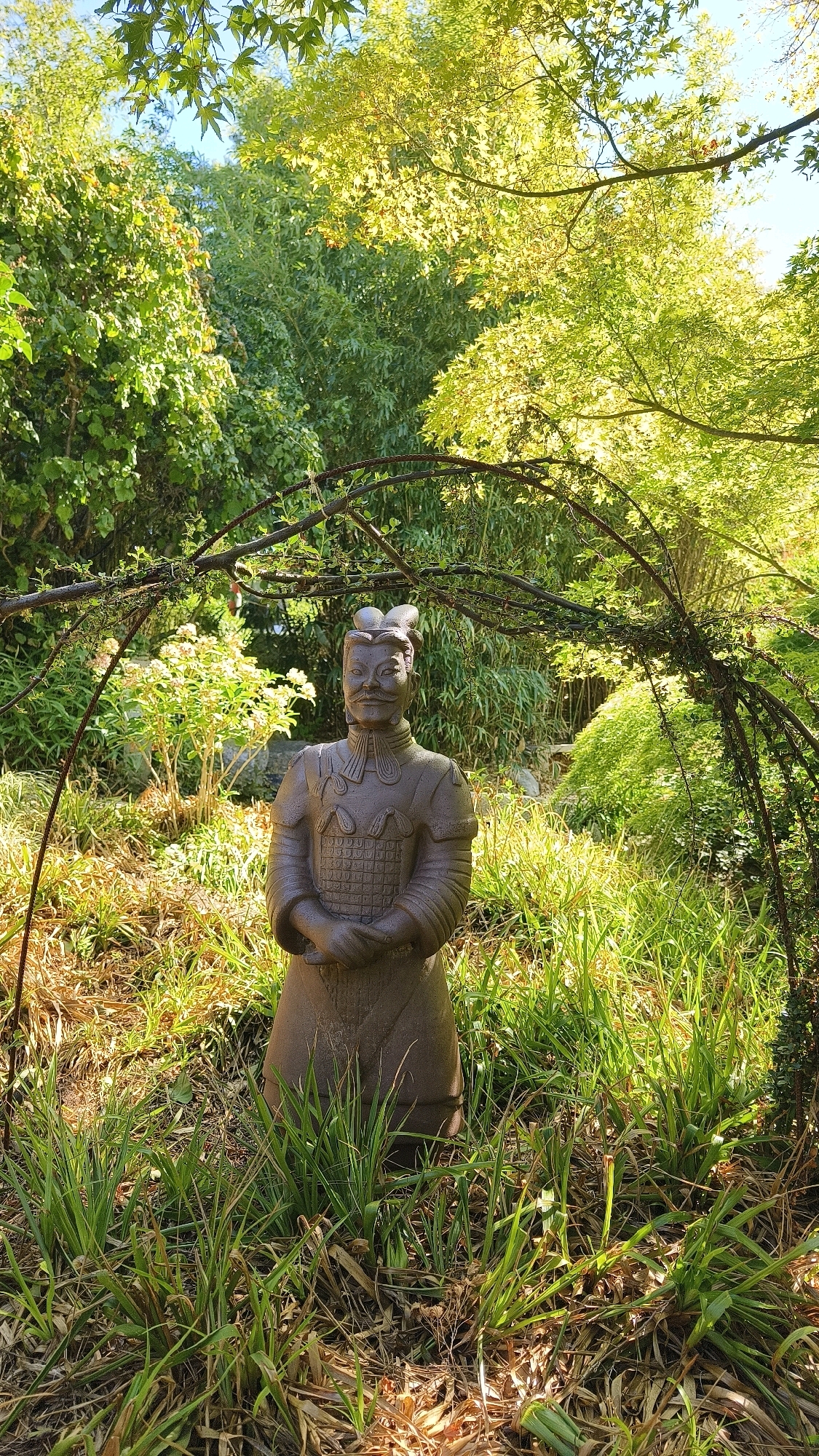 The image shows a statue of a Terracotta Warrior in a garden setting in Langenlois, Austria. The statue is made of a dark, weathered material and stands in the center of the frame, partially obscured by a decorative archway of branches and vines. The warrior is depicted in traditional armor, with its hands clasped in front of its body. The surrounding environment is lush and green, with various trees, shrubs, and tall grasses. Sunlight filters through the leaves of the trees, creating dappled light on the ground and the statue. In the background, there are more dense trees and foliage, suggesting a natural or park-like setting. The sky is visible through the canopy of leaves, appearing blue and clear.