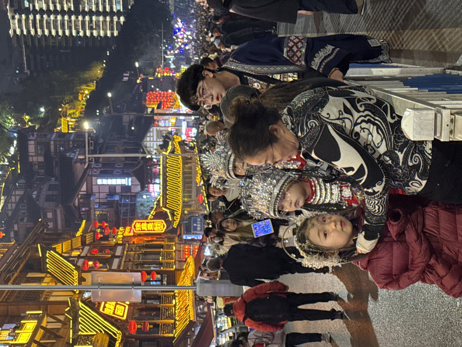 This image depicts a crowded nighttime street scene in Yuzhong District, China, characterized by vibrant artificial illumination. In the foreground, from left to right, a young girl with golden glitter on her face and an elaborate silver beaded headpiece wears a maroon puffer jacket and looks directly at the camera. Next to her, an adult East Asian woman with glasses and a black and white patterned long-sleeved shirt is leaning over a metal railing, her right hand near the young girl's head, appearing to adjust or tend to her. To the right of the woman, another young child, wearing a reflective silver headpiece and traditional-style clothing, looks straight ahead with a slightly downturned expression. A young East Asian man with dark hair and glasses, wearing a dark blue and white patterned tunic, stands behind the adult woman and children, observing the scene to his right.
The background is densely populated with numerous individuals walking in various directions. Traditional multi-story buildings, adorned with intricate wooden structures and multiple red lanterns, are heavily outlined and illuminated with yellow neon lights, creating a festive atmosphere. Some signage in Chinese characters is visible on these buildings. Beyond the immediate street level, additional buildings ascend a steep hillside, also illuminated, culminating in a taller, more modern high-rise building visible on the far right. The ground surface is dark pavement, reflecting some of the overhead lights.