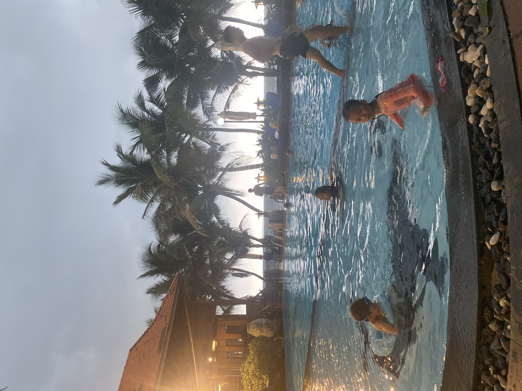 This image captures a vibrant scene at a resort swimming pool during late afternoon or early evening, likely around sunset, in Phu Thuy Ward, Vietnam. The sun, low on the horizon, casts a warm glow, creating silhouettes of numerous tall palm trees that frame the pool and resort buildings.
In the foreground and mid-ground, several individuals are enjoying the blue waters of the swimming pool. A person, possibly an adult or older child, stands partially submerged at the pool's edge, wearing dark shorts and blue arm floats. To their right, a young child in a pink full-body swimsuit stands in the water, looking towards the viewer with a faint smile. Further away in the pool, another child is visible wearing goggles, and other figures are present, suggesting a lively environment. A pair of pink swim goggles rests on the pebbled border of the pool near the right edge.
Beyond the pool, resort structures with distinctive red-tiled roofs are visible amidst the dense palm foliage. Artificial lights from these buildings are starting to glow, enhancing the twilight atmosphere. In the distant background, a bright horizon suggests the proximity of the ocean, with faint shapes of beach chairs and umbrellas along what appears to be a shoreline or an elevated deck. The sky is bright but hazy or overcast, contributing to the soft, warm lighting. The overall impression is one of leisure and relaxation in a tropical, coastal setting.