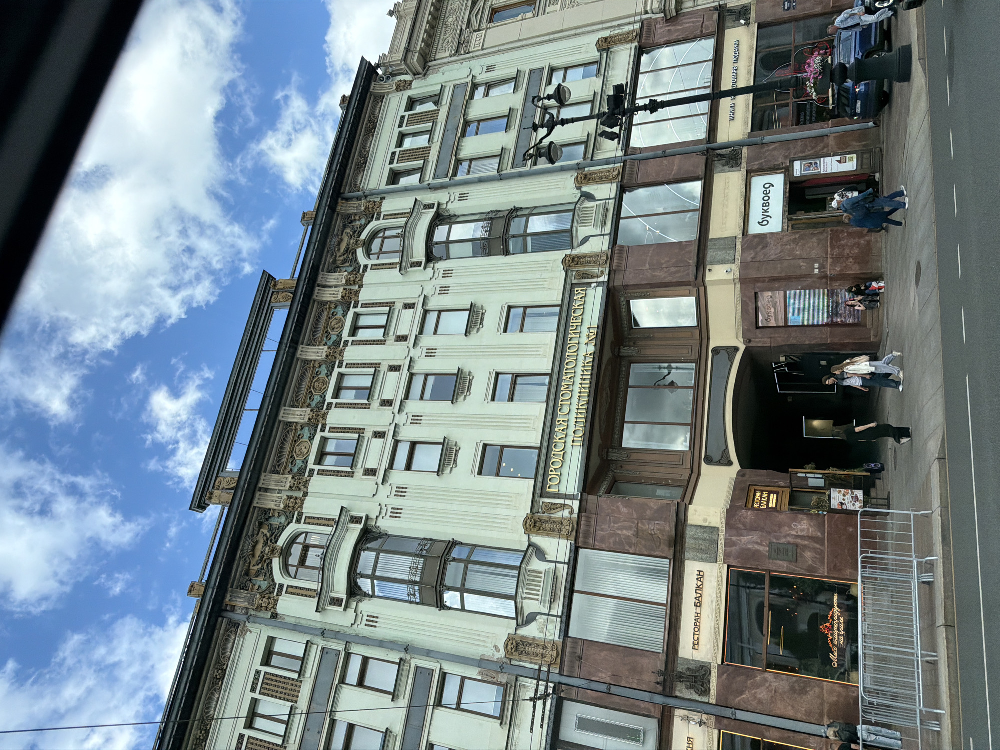 The image shows a street view of an ornate, light green building with ornate architectural details and gold lettering above the entrance. The building appears to be a commercial establishment, with signs for "ГОРДСКАЯ СТОМАТОЛОГИЧЕСКАЯ ПОЛИКЛИНИКА №1" (City Dental Clinic No. 1), "РЕСТОРАН БАЛКАH" (Restaurant Balkan), and "Буквоед" (Bukvoed, a bookstore). Several people are walking on the sidewalk in front of the building, and a dark blue car is parked near the entrance to the bookstore. The sky is blue with scattered white clouds. The architecture suggests the building is located in Saint Petersburg, Russia, likely in a historic district due to its elaborate design.