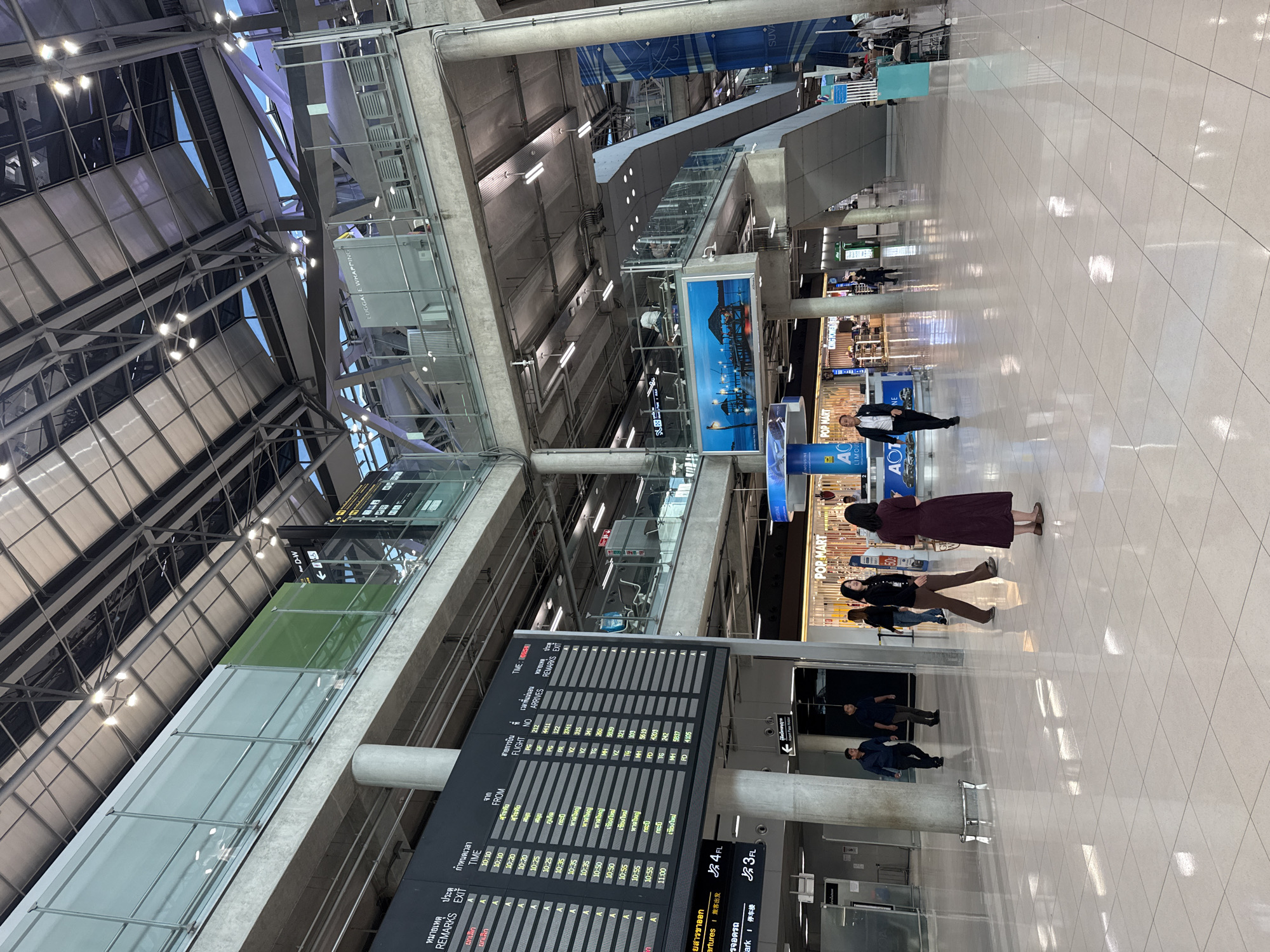 The image shows a wide, brightly lit interior of a modern airport terminal. The architecture features high ceilings with exposed metal beams and a pitched roof covered in what appears to be metallic panels. Large concrete columns support the upper levels. On the left, a large departure/arrival information board displays flight details in Thai and English. Several people are walking through the terminal. In the foreground, a woman in a maroon dress is walking away from the camera. To her left, a man in a black jacket and brown pants is walking towards the camera, followed by another man in a dark blue shirt and black pants. Further into the terminal, there are shops visible, including one with a "POP MART" sign. The floor is tiled with a reflective white surface. The overall scene conveys a sense of busy airport activity in a spacious and contemporary environment, likely Suvarnabhumi Airport, located in Nong Prue Subdistrict, Thailand.
