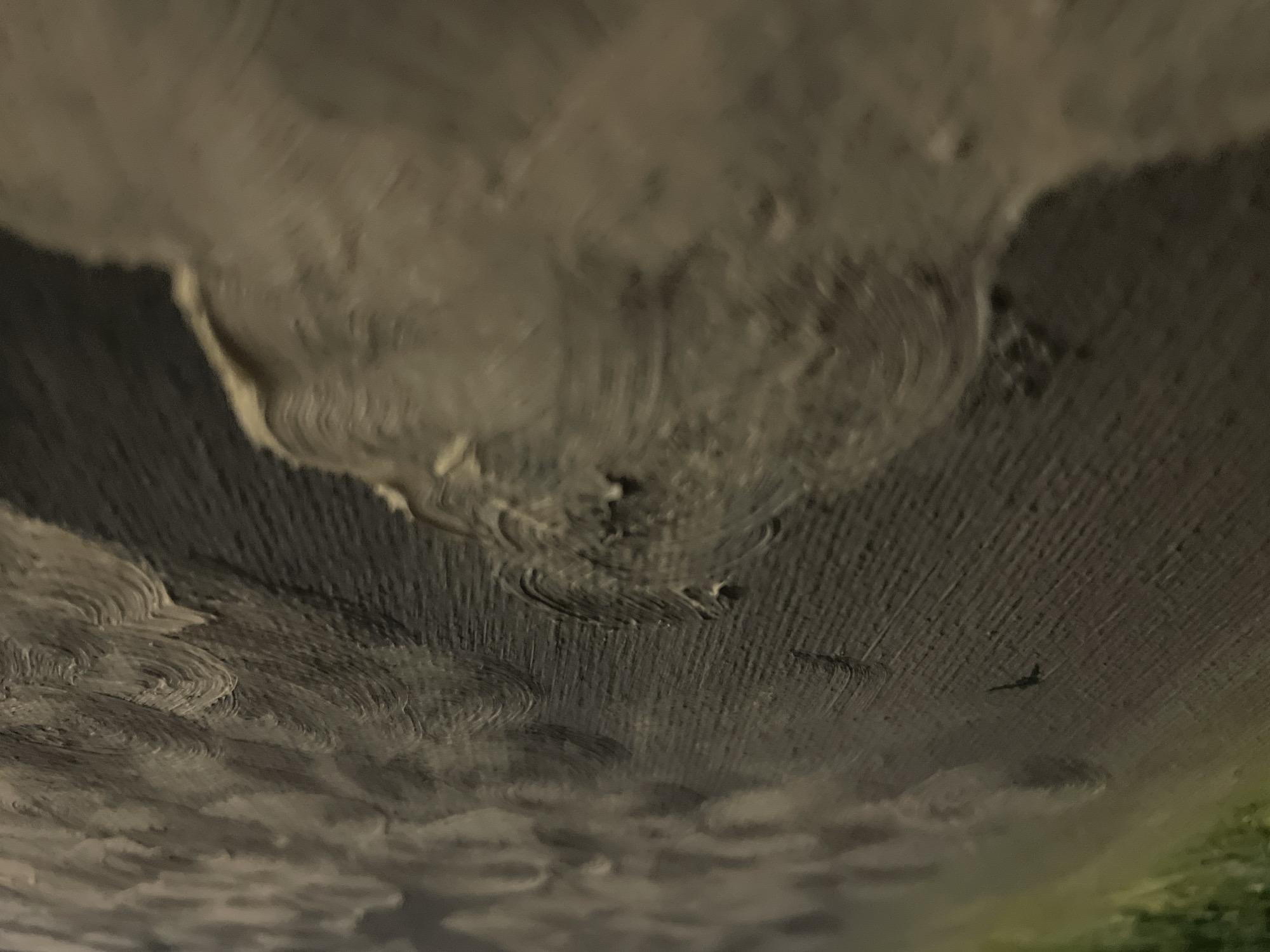 The image depicts an extreme close-up of what appears to be an oil painting, with thick, swirling brushstrokes of grey and white. The texture is highly visible, suggesting impasto technique. At the bottom right corner, there is a hint of green. A small dark shape, possibly a bird, is seen in the center of the image. The lighting is dim and uneven, highlighting the texture of the paint. There is no information to determine that this image was taken in Bac Nha Trang Ward, Vietnam.
