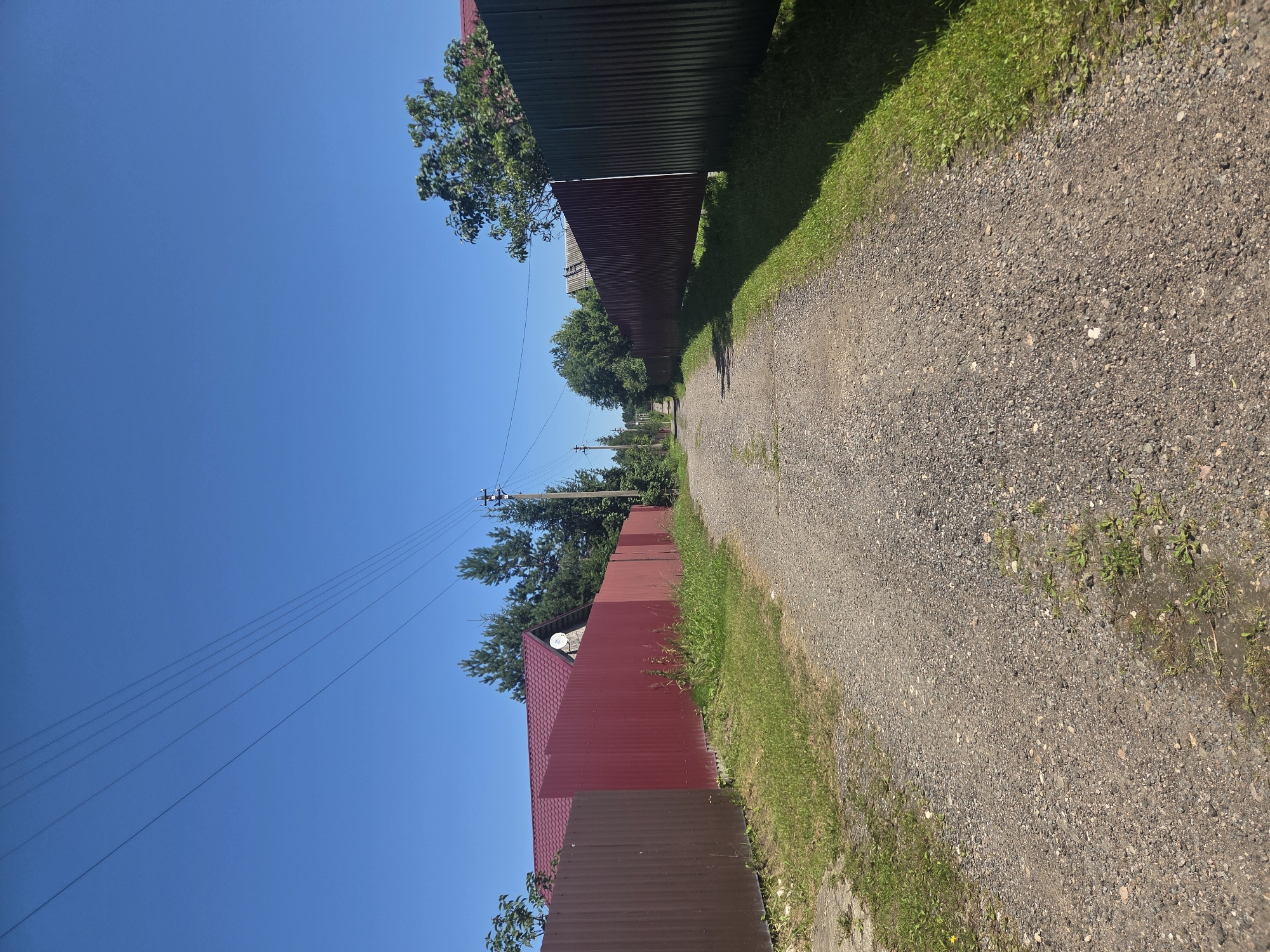 A gravel road winds through a rural area with fences on both sides, under a clear blue sky. On the left, a red metal fence runs alongside the road, with a glimpse of a house with a red roof and a satellite dish behind it. On the right, a darker metal fence, possibly brown or dark green, also borders the road. Trees are visible beyond the fences on both sides. Telephone wires are strung across the sky overhead. The overall impression is a quiet, sun-drenched day in a suburban or rural setting.