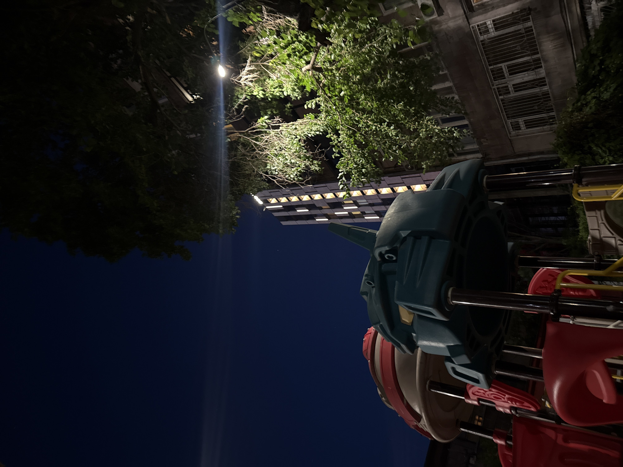 A low-angle, nighttime shot captures a section of a playground and a residential building in Yerevan, Armenia. The playground equipment, primarily red and teal plastic, is prominently featured in the foreground. Above the playground, a tall, dark residential building with illuminated windows rises into the night sky. Lush green trees with dense foliage partially obscure the building and overhang the scene, with a streetlamp casting a bright light visible among the branches. The overall atmosphere is dimly lit, with the dark blue sky serving as a backdrop.