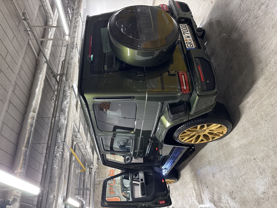 A customized Brabus G-Wagen SUV, finished in a dark metallic olive-green paint with matte gold multi-spoke wheels, is positioned within an indoor car detailing facility or garage. The vehicle's surface exhibits water droplets, suggesting it has been recently washed or is in the process. The driver's side front door is ajar, revealing an illuminated blue step and a light-colored interior with visible seating. The rear features "Brabus" badging on the tailgate and a matching spare tire cover, also branded "Brabus." The Polish license plate reads "GR W7000." The environment includes a wet, grey concrete floor, textured concrete walls, and an industrial ceiling with exposed, insulated ventilation ducts and overhead fluorescent strip lighting. A partial word, possibly "PILKO," is visible on the wall through the open door. No individuals are present in the scene. Based on the license plate and the request, the location is inferred to be an indoor automotive facility in the city of Warsaw, Poland.