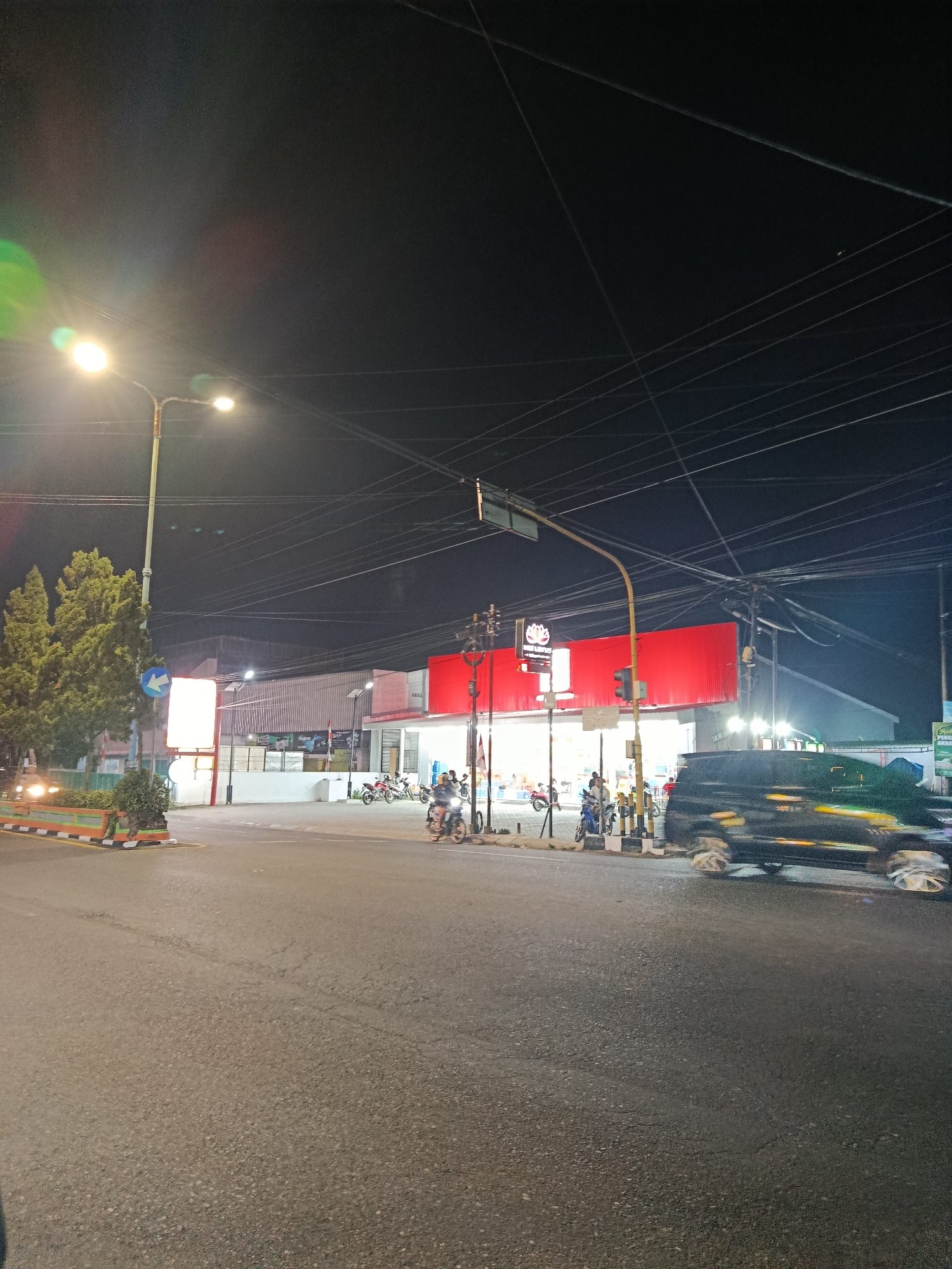 The image captures a nighttime street scene in Mlipak, Indonesia. A bright street lamp illuminates the foreground and casts a green and red lens flare onto the dark sky, which is crisscrossed with numerous electrical wires. To the left, a row of green cypress trees stands beside a white rectangular sign with a blue arrow pointing left. Further into the scene, a long building with a red facade and white accents stretches horizontally. The building appears to be a commercial establishment, possibly a supermarket, with several people and motorcycles visible in front. A black SUV is in motion on the right side of the road, its wheels blurred by the camera's exposure. The asphalt road is wet and reflects the ambient light.