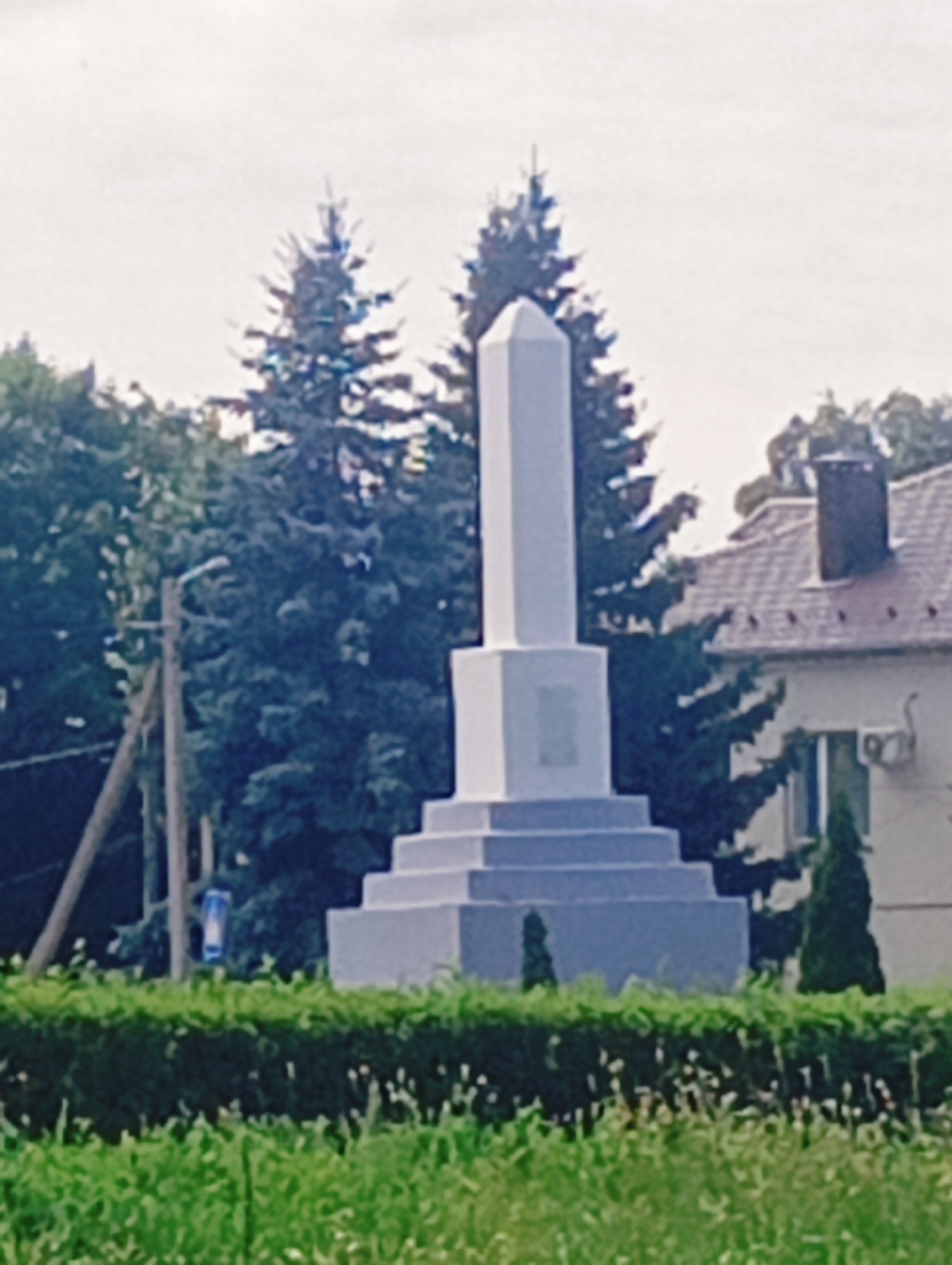 This media file depicts a monument in an outdoor setting, likely a public space such as a park or town square, in Chervonaya Slabada, Belarus. The monument is a tall, obelisk-shaped structure with a stepped base, painted a light color, possibly white or light grey. It stands prominently in the center of the frame.
Behind the monument, a dense stand of dark green coniferous trees forms a natural backdrop. To the right of the monument, a building with a reddish-brown tiled roof and a brick chimney is visible, suggesting a residential or communal area. A window and an air conditioning unit are discernible on the side of the building.
In the foreground, lush green grass and low-lying bushes with small white flowers create a natural border. A wooden utility pole with wires and a blue sign is situated to the left of the monument, adding a touch of everyday infrastructure to the scene.
The weather appears to be overcast, with a bright, diffused sky, suggesting it is daytime. There are no people visible in the image. The overall mood is serene and possibly commemorative, given the nature of the monument. No visible text is legible within the frame.