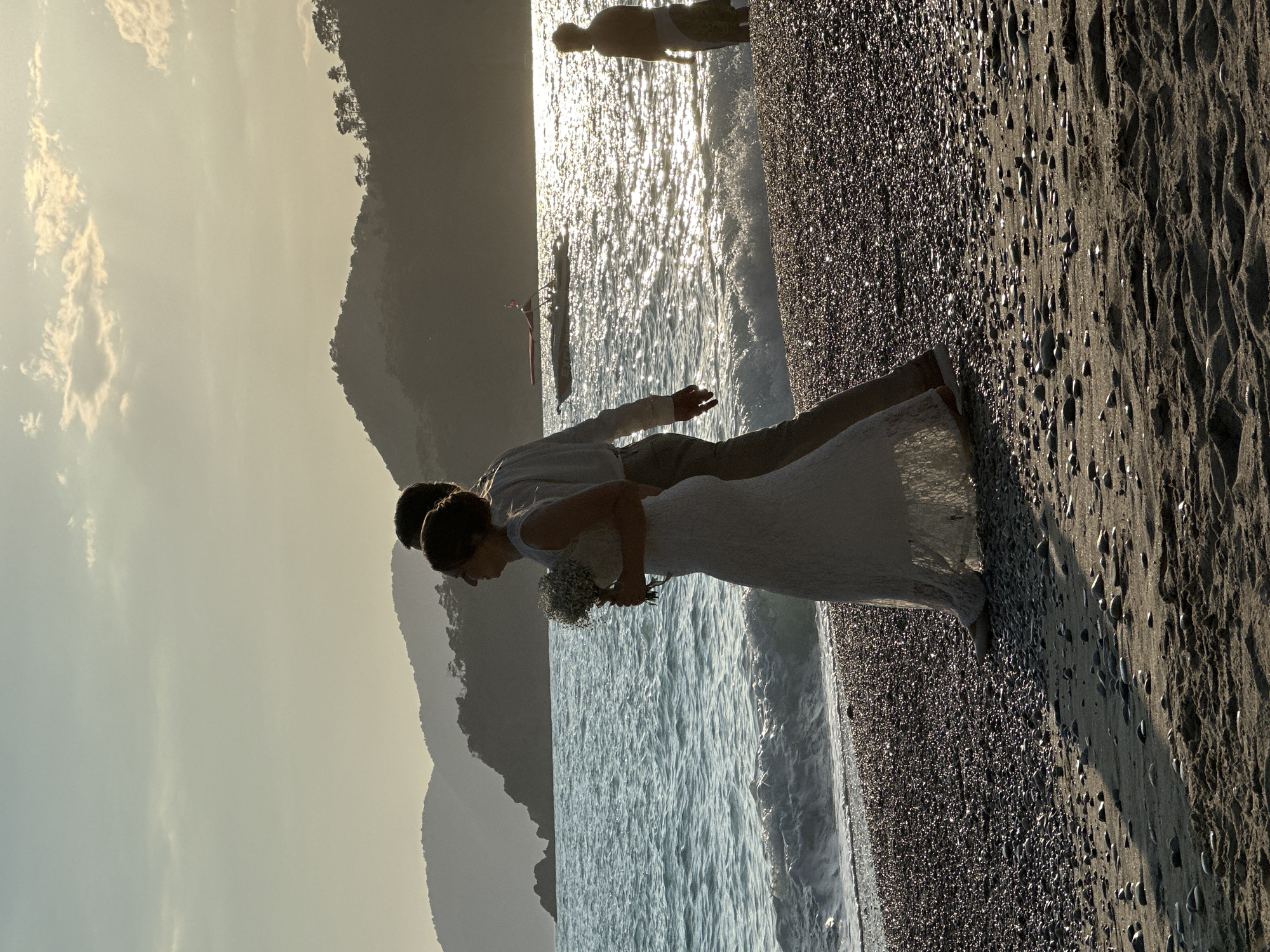 A couple is standing on a pebble beach in Fethiye, Turkey. The woman is wearing a white dress and holding a bouquet of flowers. The man is wearing a light-colored shirt and pants. They are standing near the water's edge, with the ocean in front of them and mountains in the background. The sky is overcast and a boat can be seen in the background. There is another person in the background as well.
