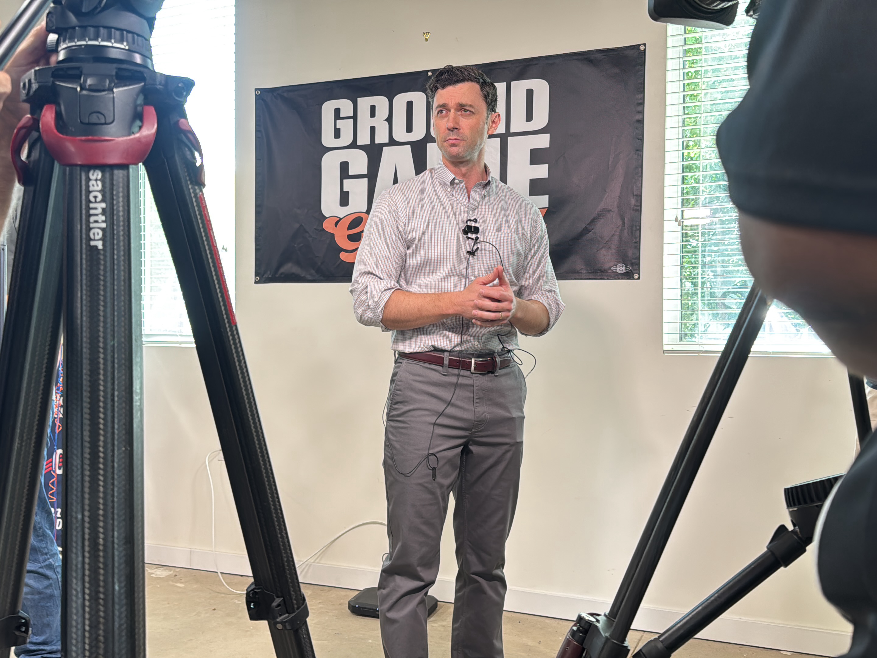 The image depicts a man standing indoors, apparently addressing a camera. He is fair-skinned, dressed in a button-down shirt and grey trousers with a brown belt. He has a small microphone clipped to his shirt. The backdrop behind him is a banner with the words "GROUND GAME" in large letters. The setting seems to be a room in a building, with two windows visible. A camera tripod is present on each side of the man, suggesting he is being filmed. The location is Decatur, United States.
