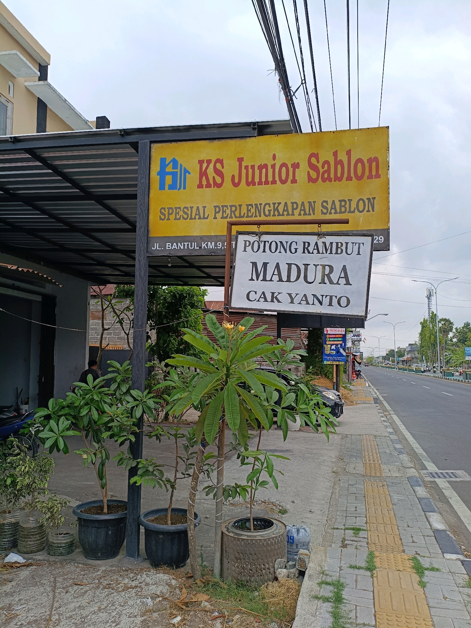 The image shows a street scene in Sewon, Indonesia. The primary focus is a yellow sign with blue and red text that reads "KS Junior Sablon SPESIAL PERLENGKAPAN SABLON JL. BANTUL KM.9, T". Below this, a white sign with black text reads "POTONG RAMBUT MADURA CAK YANTO". In the foreground, several potted plants are arranged on the ground. To the right, a sidewalk with tactile paving is visible, leading to a road with traffic. The sky is overcast.