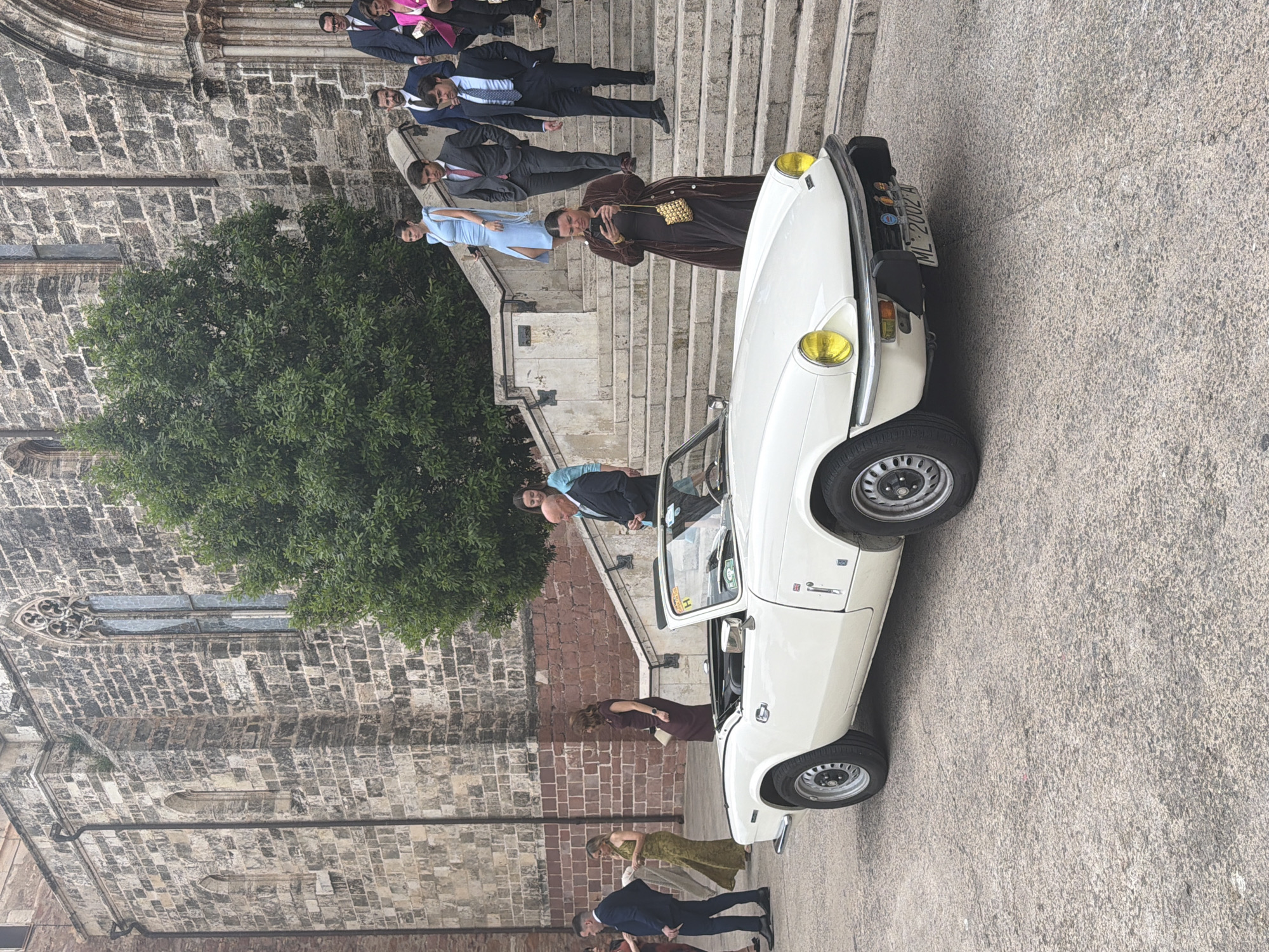 The image shows a group of people gathered outside what appears to be a church or historic building in El Puig De Santa Maria, Spain. The building is constructed of gray stone blocks. A leafy green tree is next to the building. A white vintage car, possibly a Triumph, is parked on the right side of the frame. People dressed in formal attire are on the steps leading up to the building. Several men are wearing suits, and at least one woman is wearing a light blue dress. One person is dressed in a brown robe. A license plate is visible on the car. The ground appears to be concrete or paving stones.