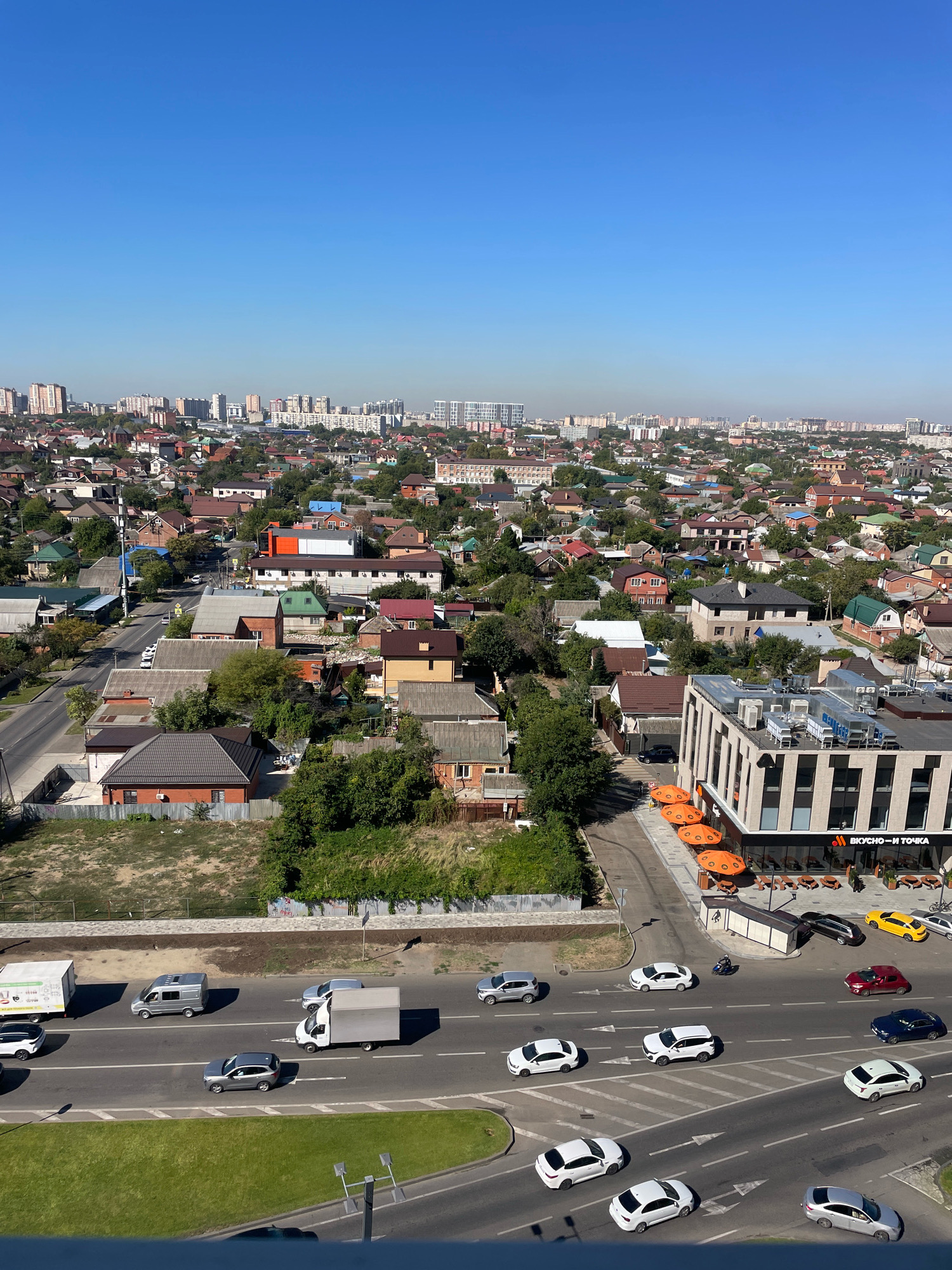 This is an aerial, full-frame, eye-level, daylight, vertical shot of a residential area in Krasnodar, Russia, with a modern commercial building in the foreground. The scene is dominated by a clear blue sky and a dense urban landscape of low-rise homes with red and brown tiled roofs, interspersed with trees. In the mid-ground, a road runs through the neighborhood, carrying a moderate amount of traffic, including cars, vans, and trucks. A white van with a cargo box is driving on the right side of the road, with a silver minivan and a white SUV also visible. Further down the road, a white truck is seen, and several other cars are present.
To the right of the main road, a multi-story commercial building with a modern facade, featuring large windows and outdoor seating with orange umbrellas, is situated. A sign on the building displays the Russian text "ВКУСНО — И ТОЧКА," indicating a fast-food establishment. A motorcycle with a rider is navigating a street in front of this building, and several cars are parked or moving nearby, including a black sedan, a yellow hatchback, and a red sedan.
The foreground of the image shows a grassy embankment sloping down towards the road, with some paved areas and streetlights visible. The overall impression is a snapshot of a typical day in a developed urban environment, where residential areas coexist with commercial activity and transportation infrastructure. There are no visible people in focus or distinct interactions being captured.