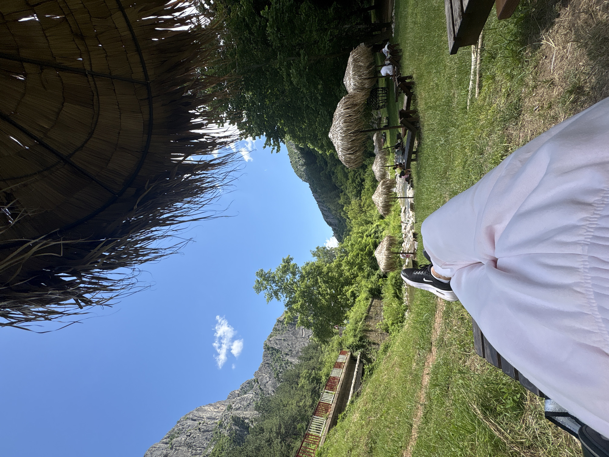 An outdoor perspective, looking up from a reclining position under a thatched umbrella, reveals a scenic mountain landscape. In the foreground, a person's legs are visible, clad in light pink trousers and black and white sneakers, resting on a wooden lounge chair. Beyond the chair, a lush green lawn stretches towards a river. Several thatched umbrellas with accompanying lounge chairs are arranged on the grassy bank, with a few people seated. A red and white railing borders a concrete structure on the left, leading uphill towards a forested mountain. The sky is a vibrant blue with scattered white clouds. The scene is set in the natural park of Zgorigrad, Bulgaria, a location known for its natural beauty and recreational opportunities.