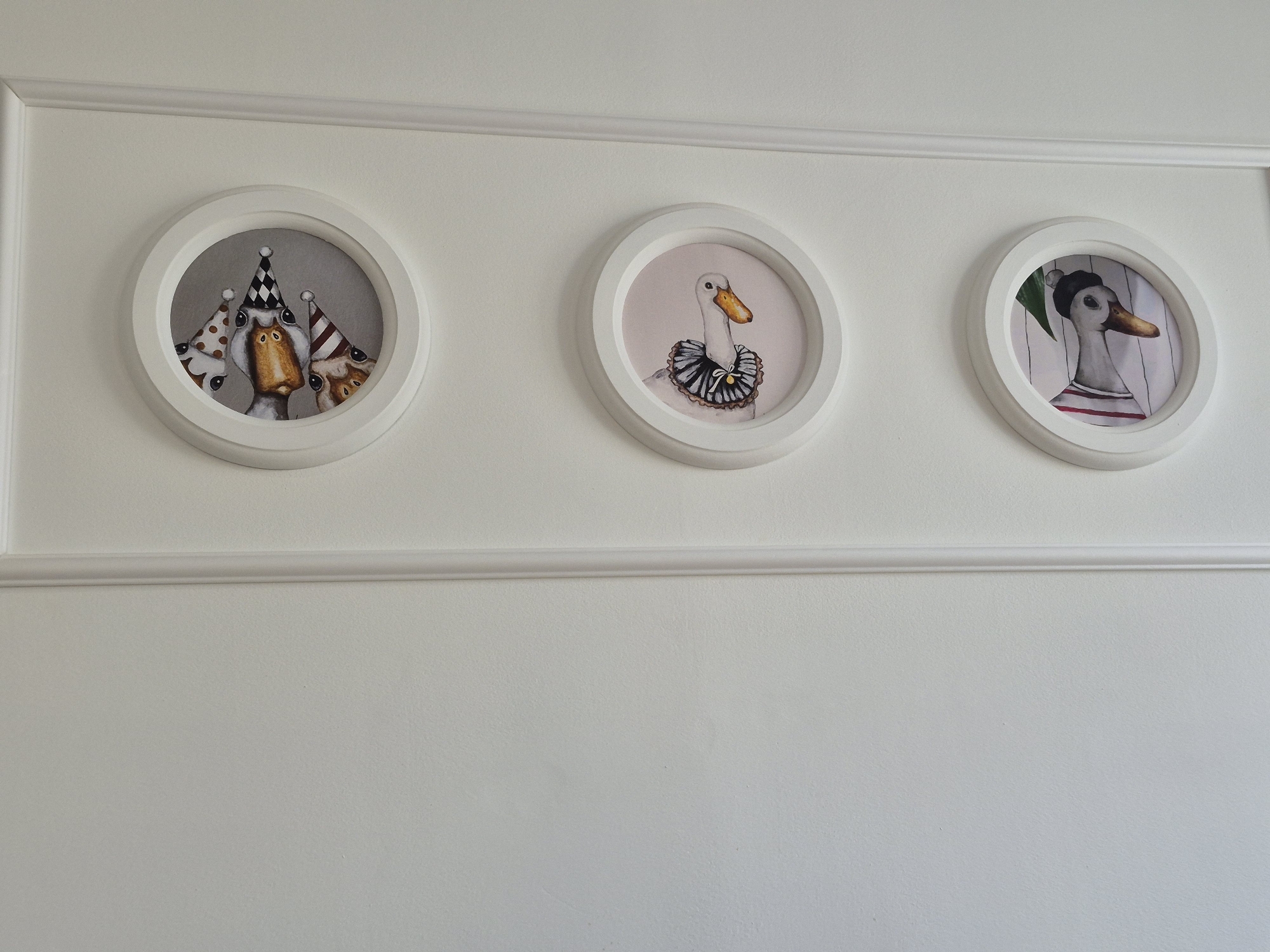 The image depicts three round, white-framed pictures hanging on a white wall with a horizontal molding running behind them. The leftmost picture contains three cartoon ducks wearing party hats. The center picture displays a cartoon duck wearing a frilly collar. The rightmost picture shows a cartoon duck wearing a sailor's hat. The setting appears to be an interior space with white walls and simple decor. There is no indication of the location being in Chaikovsky, Russia.