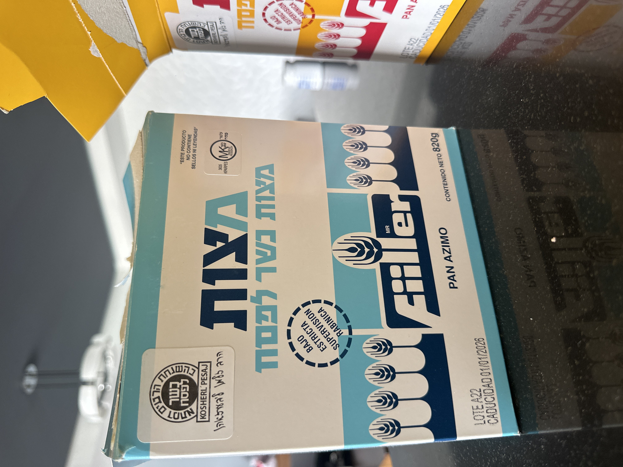 A close-up shot features a box of Filler Pan Azimo matzah on a dark surface, likely a countertop. The box has Hebrew and Spanish text, with symbols indicating it is Kosher for Passover. The expiration date printed on the box is January 1, 2026. Another box with similar labeling can be partially seen in the background. The overall environment appears to be a kitchen or food preparation area. The city of Huixquilucan de Degollado, Mexico, is not directly verifiable from the image content.