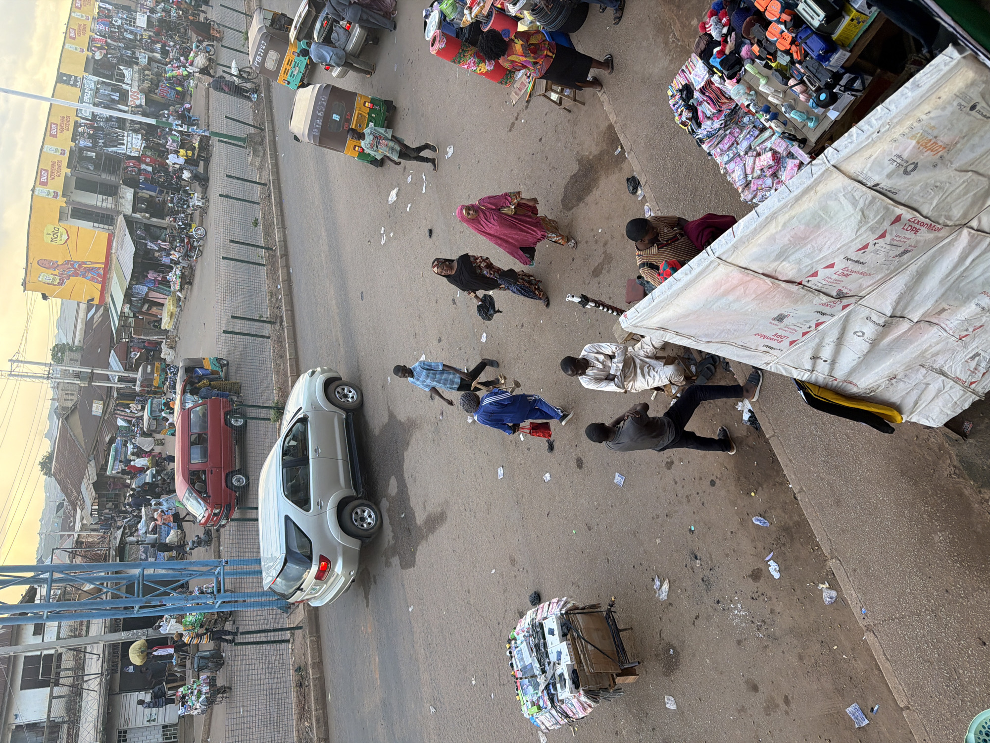 The image depicts a bustling street scene in Bukuru, Nigeria, likely a market area. The foreground shows a street lined with various stalls and vendors. In the middle ground, a silver Toyota Fortuner SUV is parked on the right side of the road. Several people are visible on the street, some walking, others engaged in conversation or attending to stalls. A street vendor is seen pushing a cart laden with goods. In the background, a row of shops with various merchandise displayed outside line the street. The architecture suggests a typical urban environment in Nigeria, with a mix of single-story and multi-story buildings. The overall atmosphere is that of a busy commercial hub.