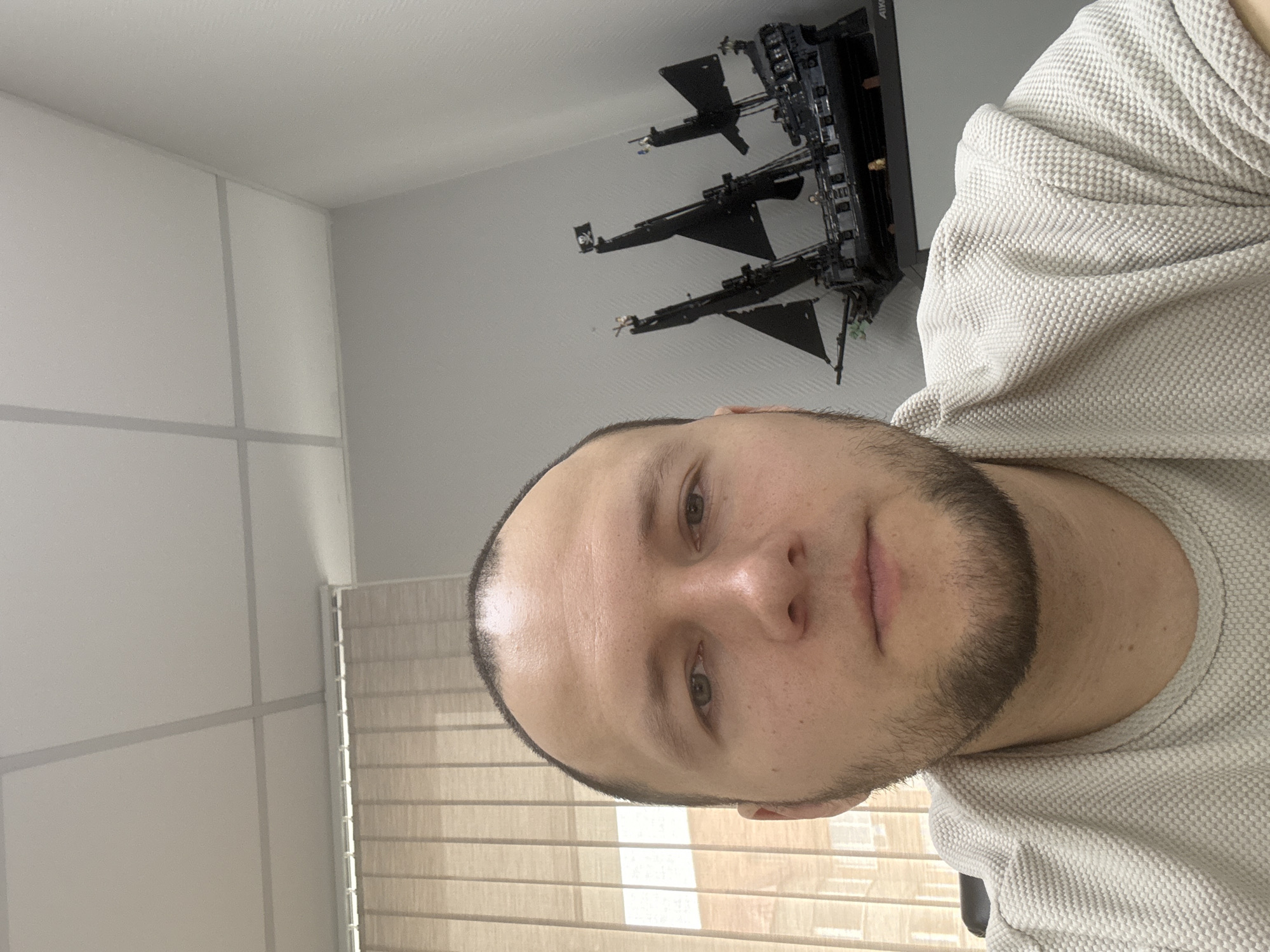 A medium shot captures a person with a shaved head and a short beard, wearing a light-colored, textured shirt. The person's gaze is directed toward the camera. Behind the person, on the wall, are mounted objects resembling model airplanes or drones. A window with closed blinds is visible to the left. The location is unknown and there is no evidence it is located in Krasnoyarsk, Russia.