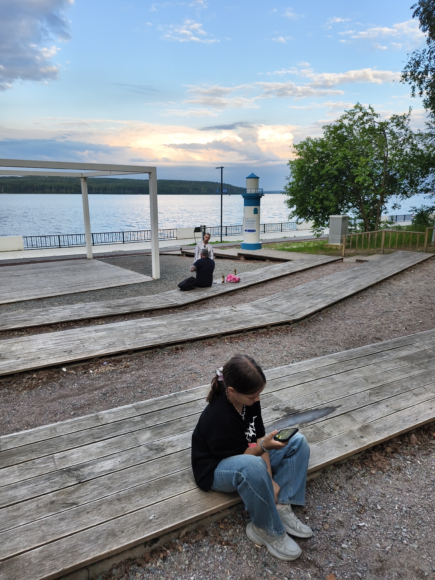 In Chaikovsky, Russia, a young girl sits on wooden bleachers, engrossed in her smartphone. She is wearing a black t-shirt, blue jeans, and grey sneakers. Her hair is pulled back in a ponytail adorned with a pink clip.
In the background, two people are seated on the ground, facing a woman standing. They appear to be engaged in a conversation or a performance. The scene is set by a lake, with a tree-lined shore visible in the distance. A small lighthouse stands near the water's edge. The sky is partly cloudy, with soft light suggesting it is either early morning or late afternoon. The overall mood is peaceful and relaxed. The weather appears to be pleasant.