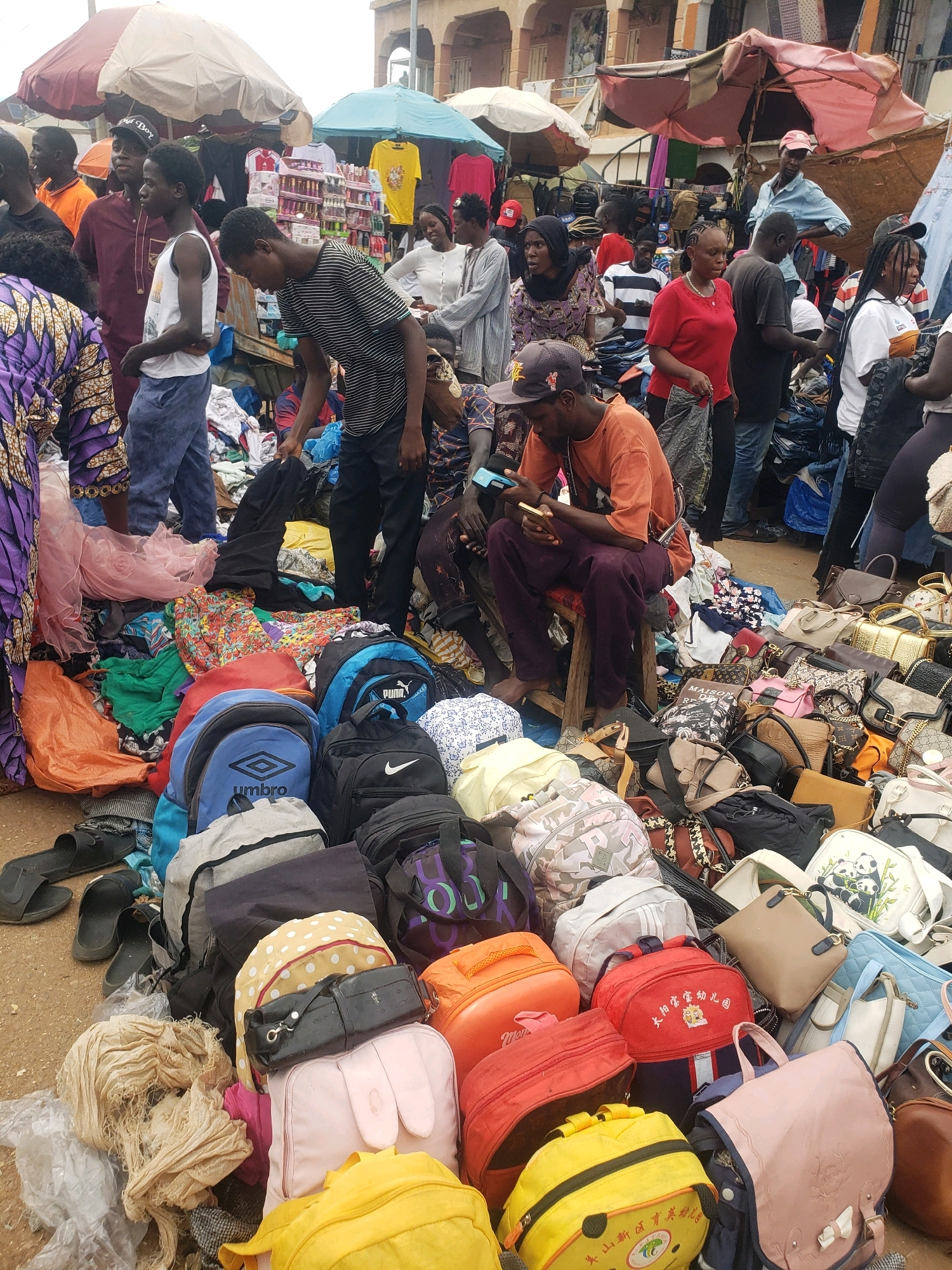 A street market scene in Sabon Tasha, Nigeria, is depicted. In the foreground, an assortment of backpacks, handbags, and other bags are piled on the ground, suggesting a retail or resale environment. Some bags are colorful and patterned, while others are more neutral in tone. Among the bags, loose clothing items and fabrics are also visible. A person in an orange t-shirt and purple pants is seated on a stool, looking at a mobile phone. Behind this person, a crowd of individuals are browsing or working in the market. The background shows market stalls with various goods, including clothing and what appear to be packaged items on shelves, along with umbrellas providing shade. The overall atmosphere is bustling and indicative of a busy marketplace.