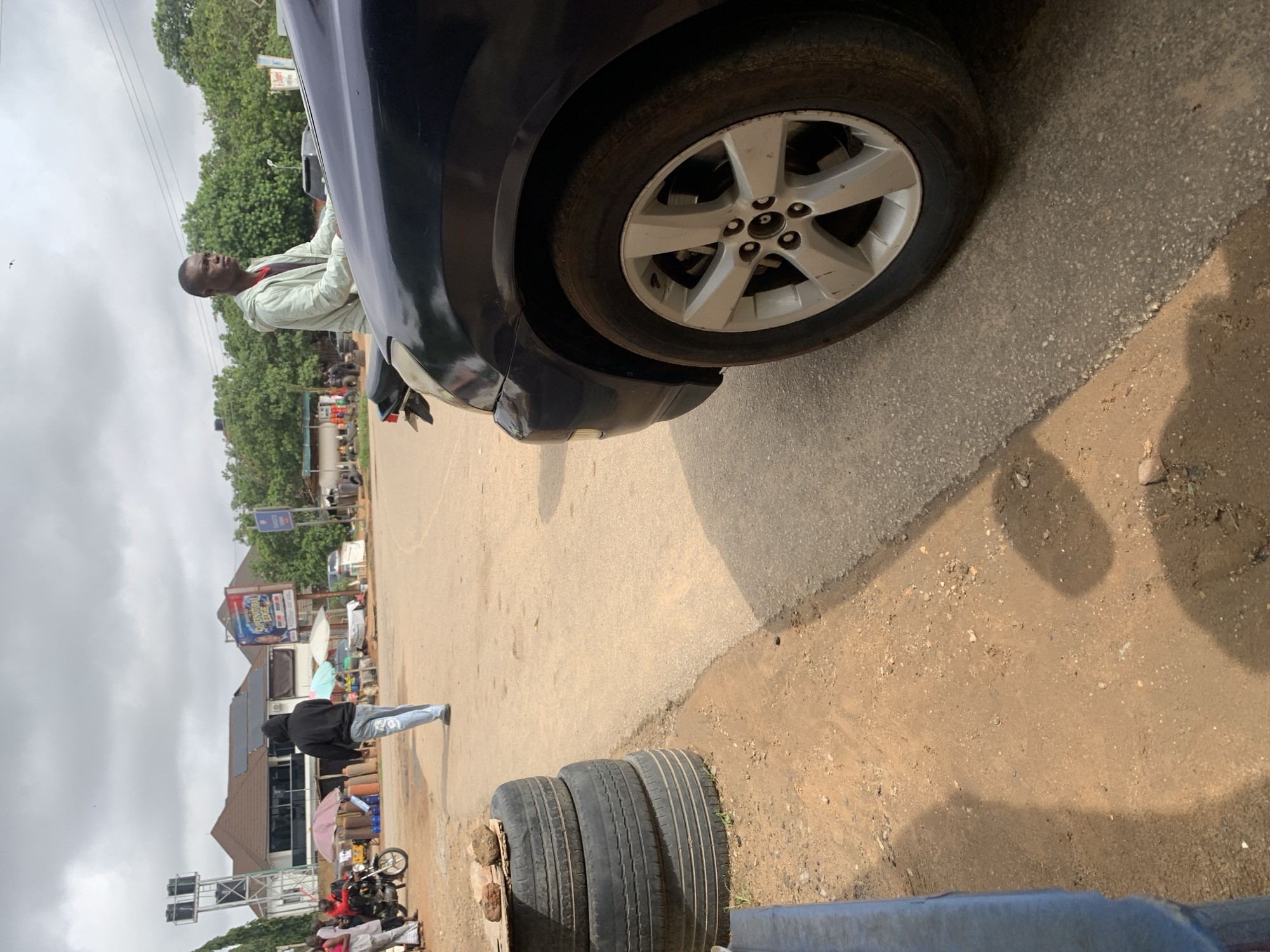 In Abuja, Nigeria, on an overcast day, a dark blue sedan is parked at an intersection. The car's front passenger-side wheel is prominently displayed in the foreground, a silver five-spoke rim contrasting with the dark tire. To the left of the vehicle, a stack of three worn tires rests on the dusty, reddish-brown ground, topped with two rocks. Beyond the tires, a person wearing a black hooded sweatshirt and blue jeans walks away from the camera, their back to the viewer. In the background, a busy street scene unfolds with various structures, including buildings with solar panels, a water tower, and market stalls with umbrellas. A billboard displaying text and images is visible on one of the buildings. Several people are gathered near a motorcycle, and another individual, dressed in a light green jacket over a red shirt, sits on what appears to be a motorcycle or scooter, looking to the right. Lush green trees line the background, adding to the urban landscape. The ground is a mix of paved road and unpaved earth.