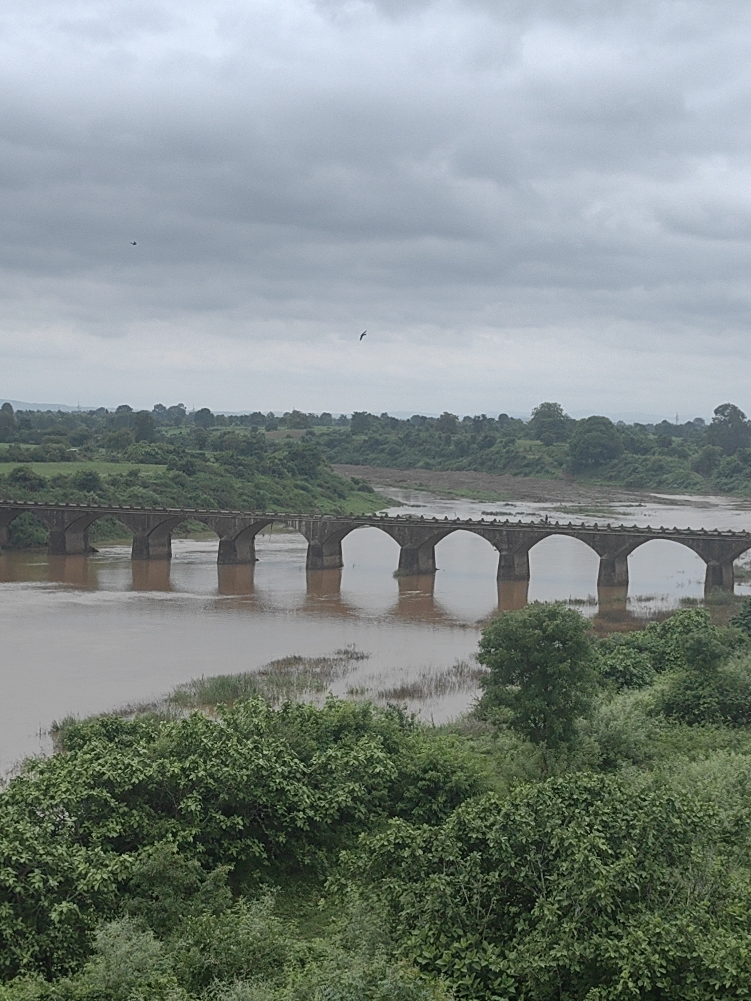 The image shows a cloudy sky over a wide, muddy river and a stone arched bridge. The bridge has multiple arches and is supported by thick pillars. The riverbanks are lined with lush green trees and vegetation. In the distance, there are rolling hills. A small bird is visible in the sky. The scene appears to be natural and rural.