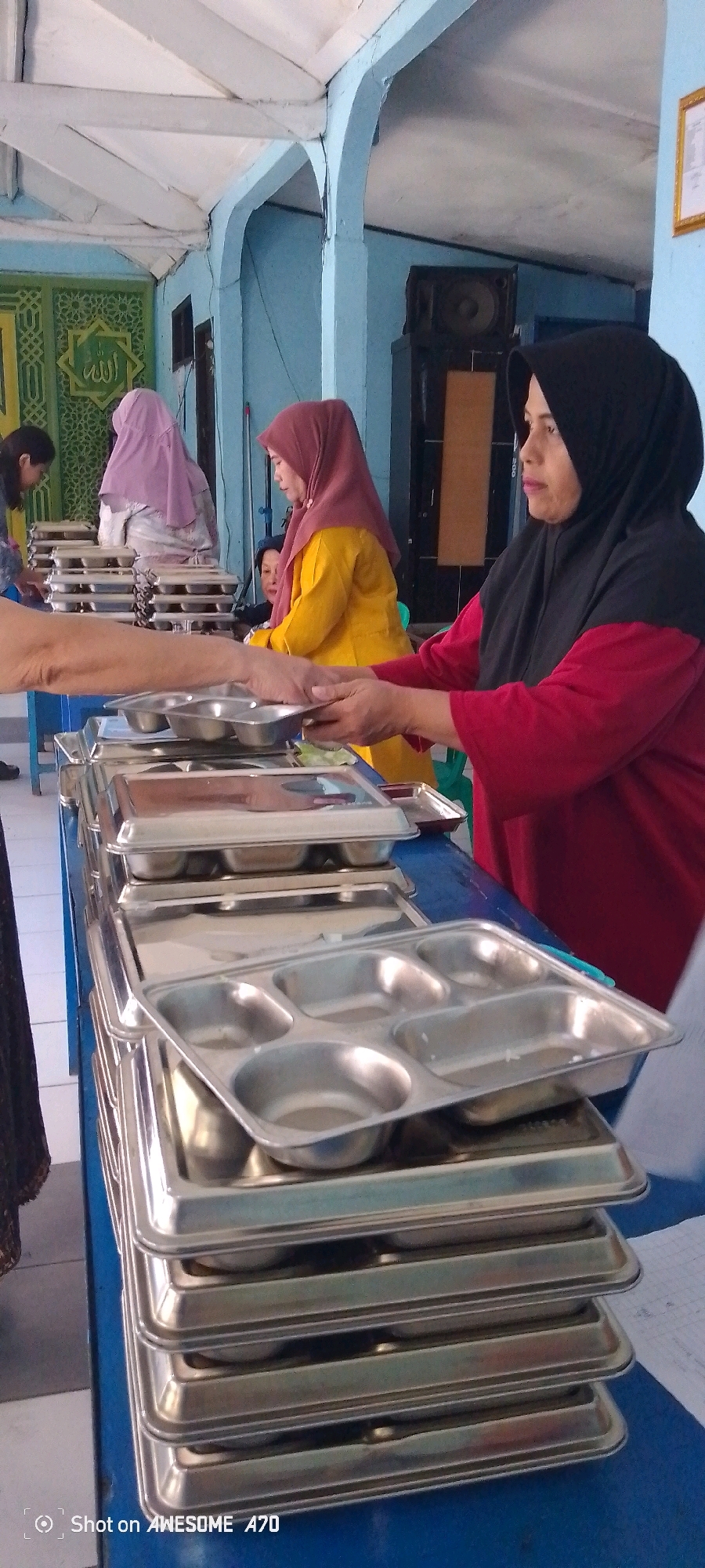 The image shows a serving line at what appears to be a community event or religious gathering in Bandung City, Indonesia. Several individuals are present, primarily women wearing hijabs. In the foreground, a stack of compartmentalized metal trays is visible, suggesting a meal is being served. One woman, wearing a red shirt and a black hijab, is holding a tray, possibly receiving food. Behind her, another woman in a yellow top and maroon hijab is also near the serving area. Further back, a woman in a light purple hijab stands behind a table with more trays. The environment seems to be an indoor or semi-outdoor space with light blue walls and a white ceiling with exposed beams. A green decorative panel with Islamic calligraphy is visible on the left wall. The overall scene depicts a communal meal service.