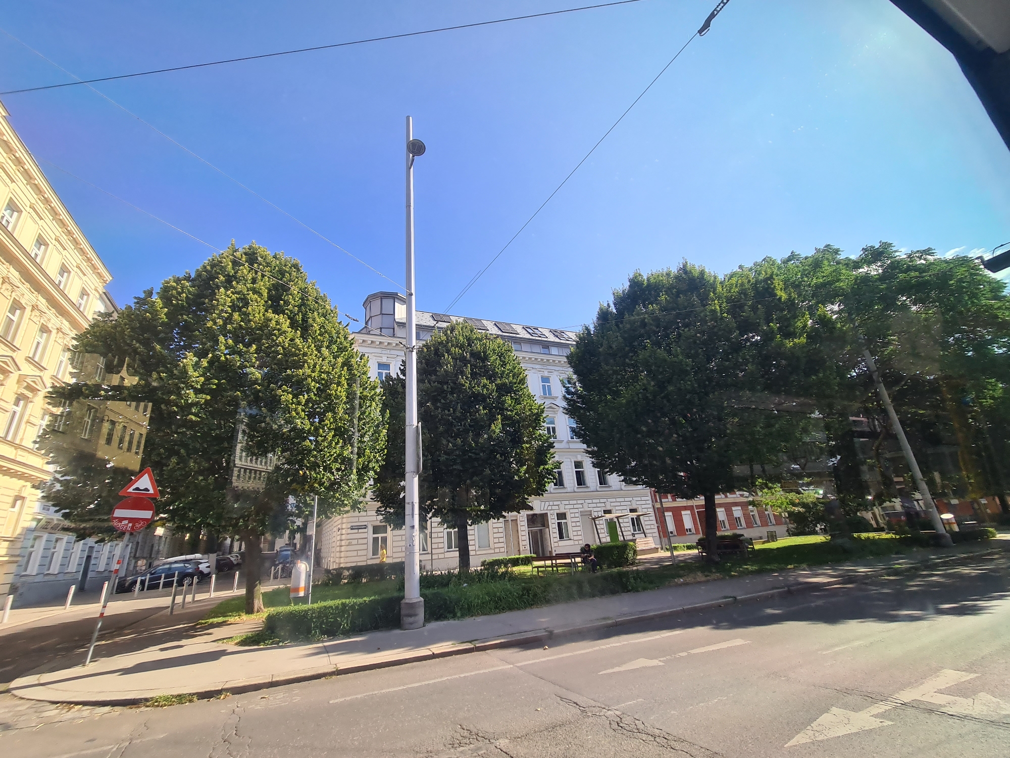 The image depicts a sunny street scene in Vienna, Austria, with buildings and trees lining the road. A light-colored building with multiple windows and ornate architectural details is visible on the left side of the image. Adjacent to it, a row of lush green trees stands tall, their branches casting shadows on the sidewalk and road. Further down the street, another building with a white facade and numerous windows can be seen, partially obscured by trees. A modern street lamp with a single light fixture stands prominently in the foreground. The road in the foreground shows signs of wear and tear with visible cracks. Traffic signs, including a triangular sign with a red border indicating a warning and a circular sign with a red border and a bicycle symbol, are positioned on the sidewalk. Several cars are parked along the street. In the middle ground, a bench is placed near a grassy area and more trees. A person is seated on the bench, facing away from the camera. The sky above is a clear, bright blue. The image appears to be taken from a moving vehicle, possibly a tram or bus, as evidenced by the reflections on the surface from which the photo was captured.