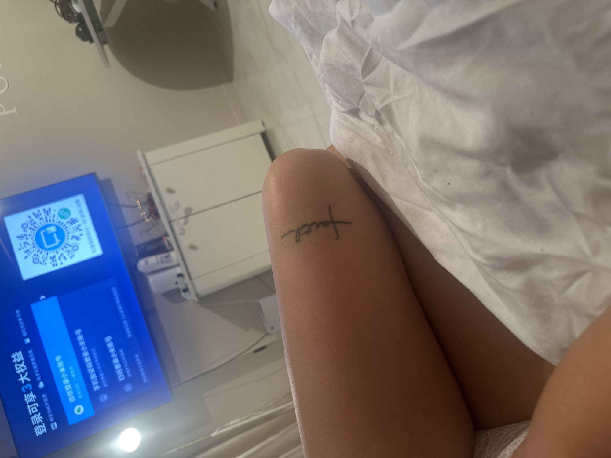 The image presents an indoor scene from a low, reclining perspective, showing a person's legs partially covered by white, crumpled bedding. A small tattoo of the word "faith" in a script font is visible on the upper thigh.
In the background, a flat-screen television is displaying a user interface predominantly in Chinese characters. The screen features a large QR code and various login options for a "小米账号" (Xiaomi account). Visible text on the TV includes "登录可享3大权益" (Login to enjoy 3 major benefits), "1080P画质同步云端着历史活动不停" (1080P picture quality sync cloud history activity non-stop), "微信登录小米账号" (WeChat login Xiaomi account), "手机验证码登录小米账号" (Mobile verification code login Xiaomi account), and "扫码登录小米帐号" (Scan code login Xiaomi account). The presence of Chinese text on the TV contradicts the given location context of Moscow, Russia.
Behind the television, a light-colored piece of furniture, possibly a cabinet or dresser, is visible. A bright light source emanates from a lower position in the room. The overall setting suggests a relaxed environment, potentially a bedroom or living room. The time of day is not explicitly indicated, with the illumination appearing to be artificial indoor lighting. No specific weather conditions or emotions are discernible.