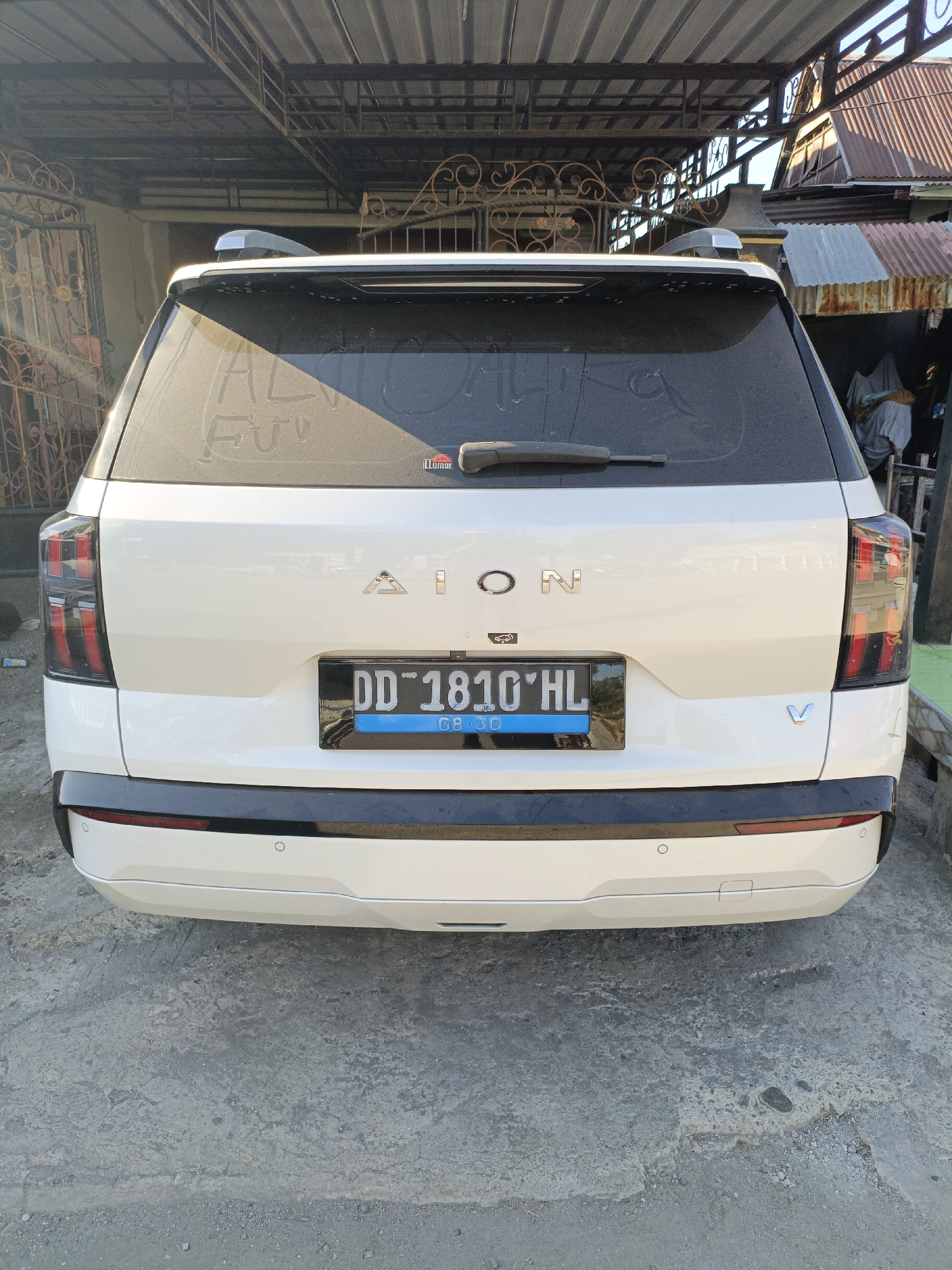 A white AION V is parked on a concrete surface under a corrugated metal roof structure. The rear of the vehicle is visible, showing its taillights, license plate which reads "DD 1810 HL", and the car's emblem. The license plate indicates it is registered in Indonesia. The environment suggests a residential area, with a gate and part of a house visible in the background. The ground is rough and appears to be a mixture of concrete and dirt. The overall scene depicts an outdoor parking area.