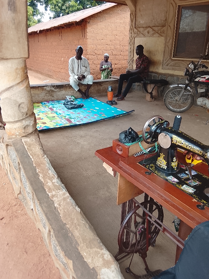 The scene depicts an outdoor covered porch area of a building, likely a residence or small business, within Akwanga, Nigeria.
In the immediate foreground, a low, rough-hewn stone and cement wall runs horizontally across the bottom of the frame. Beyond this, a reddish-brown dirt path transitions into a lighter concrete floor under the porch. A vintage black Singer sewing machine, with a wooden table surface, occupies the right foreground. It has a black spool of thread, yellow-handled scissors, a tape measure, and a small black plastic bag resting on its surface. A partial view of a human leg in dark trousers is visible next to the machine, positioned near the foot pedal.
Under the porch, a pale blue children's play mat, featuring multi-colored geometric and pictorial designs, is spread on the concrete floor. On the mat, a black bag and small items are present. An elderly man, with dark skin and a bald head, is seated barefoot on a low concrete ledge to the left of the mat. He wears a light-colored long-sleeved tunic and trousers, and faces forward. Behind him, slightly to the right, an elderly woman with dark skin stands. She is dressed in a green and white patterned wrap-around garment and a white head covering, holding an object in her hands, and appears to be looking towards the seated man. Further to the right, a younger man with dark skin is seated on a dark wooden bench. He wears a dark patterned long-sleeved shirt and dark trousers, with dark shoes visible on the ground beside his feet. He also faces towards the elderly man. A black thermos flask and a pair of dark sandals are situated on the concrete floor between the play mat and the younger man. A black motorcycle with chrome accents is parked to the right of the bench.
The back wall of the porch features decorative relief work and a window frame. In the background, beyond the porch, a reddish-brown adobe or mud brick wall with a corrugated metal roof is visible under a treeline and open sky.